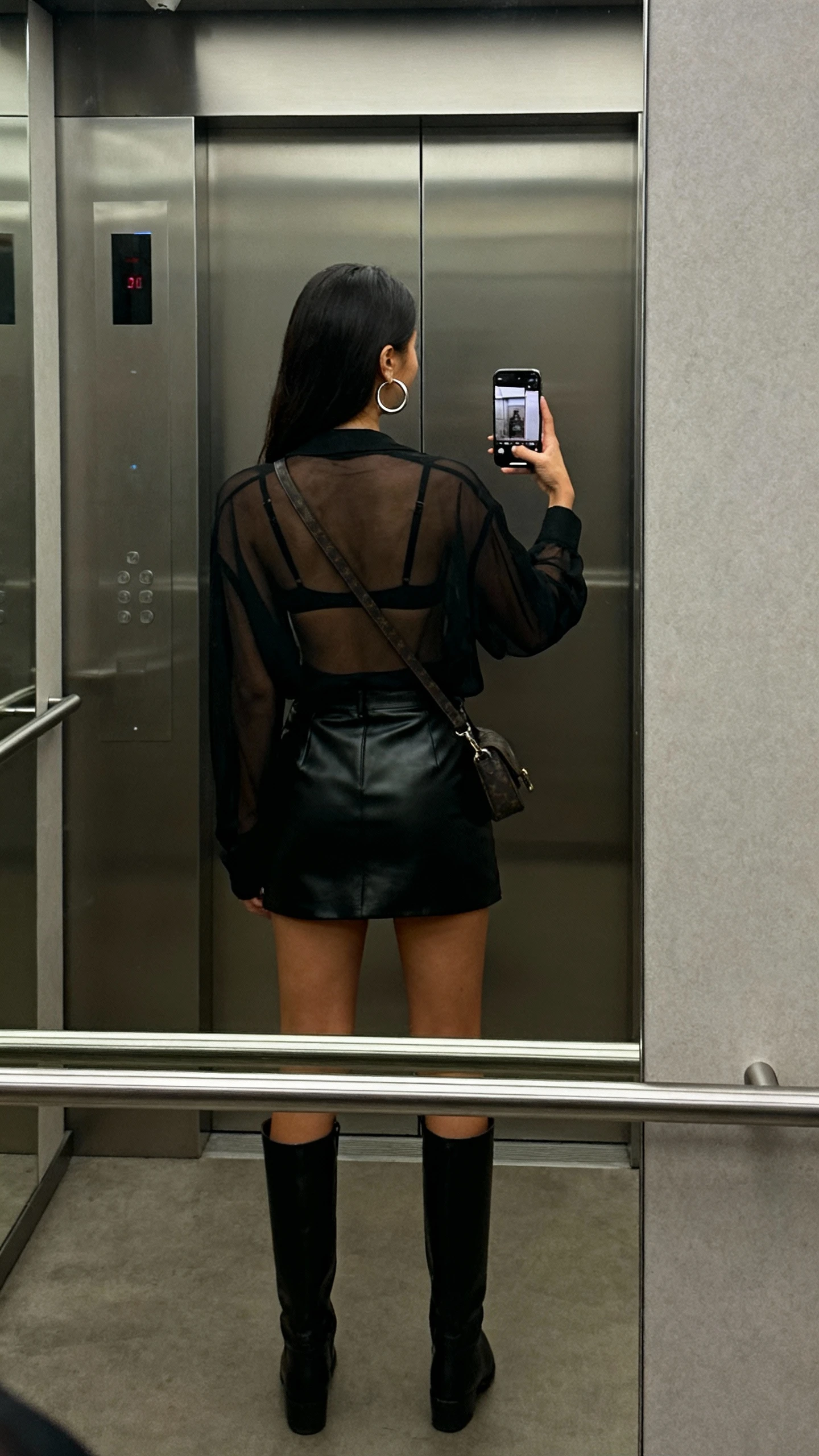 iPhone back-view selfie of a woman in a sheer black top over a bralette, leather mini skirt, knee-high boots, hoop earrings, and a crossbody bag, face not visible, elevator mirror, dim indoor lighting, natural iPhone photo quality
