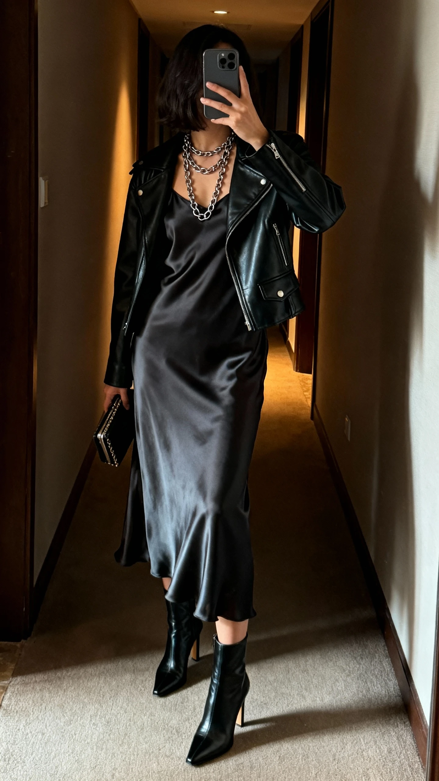 iPhone back-view selfie of a woman wearing a black leather jacket over a satin slip dress, heeled ankle boots, layered chain necklaces, small clutch, face not visible, dim hallway with warm indoor light, natural iPhone aesthetic.