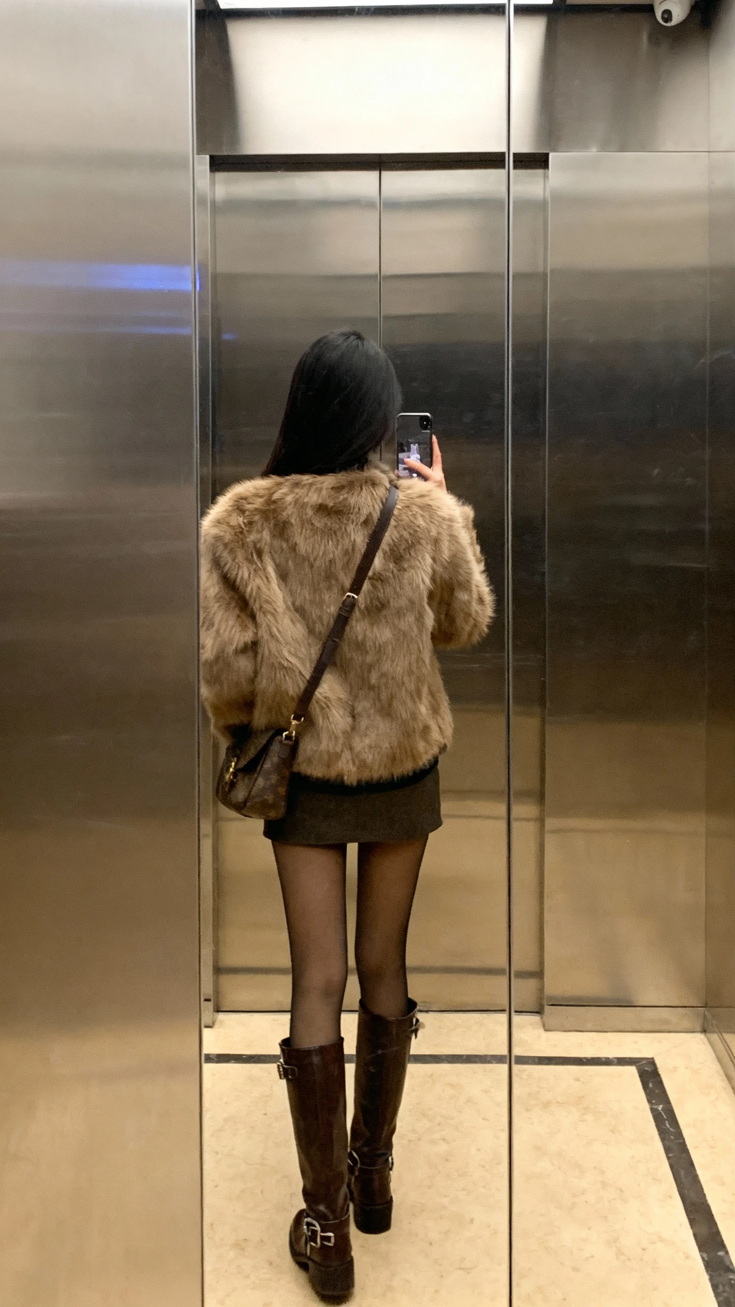 iPhone back-view selfie of a woman wearing a faux-fur jacket over a mini skirt with tall boots, tights, shoulder bag, face not visible, elevator mirror, warm indoor lighting, natural iPhone photo quality.