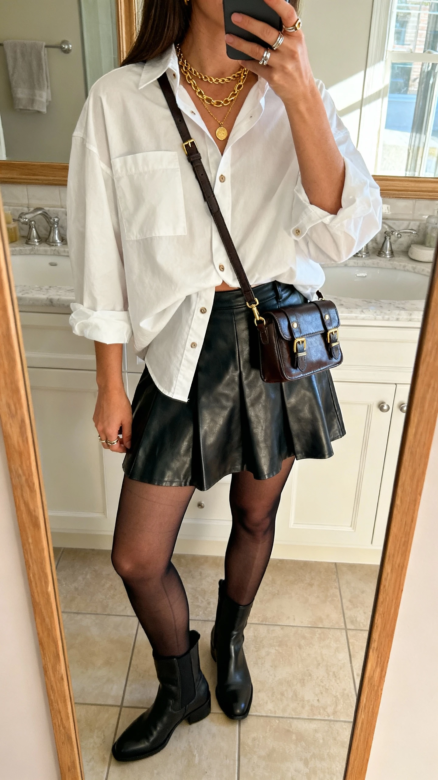 iPhone back-view selfie of a woman wearing a leather A-line mini skirt, oversized white button-down half-tucked, ankle boots, crossbody bag with hardware, layered gold necklaces, stacked rings, sheer black tights, face not visible, bathroom mirror, morning light, natural iPhone quality.