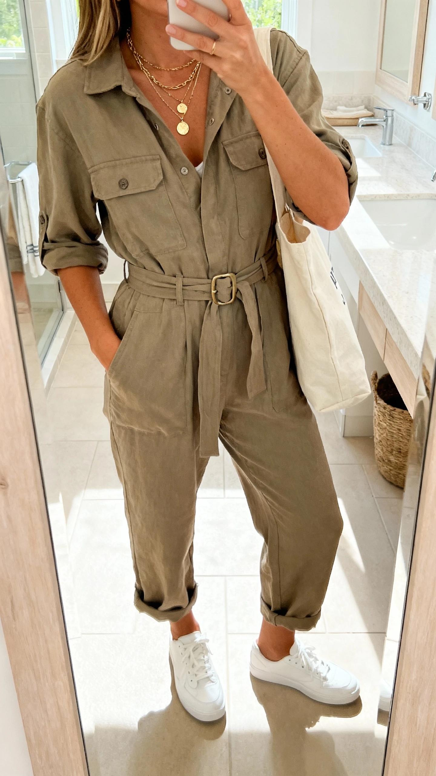 iPhone bathroom mirror selfie of a woman in a relaxed utility-style jumpsuit cinched with a belt, white sneakers, layered pendant necklaces, tote bag, face not visible, cropped at neck, bright natural light, natural iPhone aesthetic.