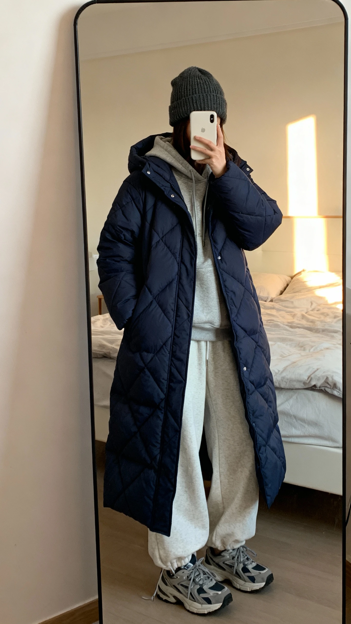 iPhone bedroom mirror selfie of a woman in a longline quilted puffer (ankle length, hooded) in deep navy, cozy hoodie, wide-leg sweatpants, chunky sneakers, knit beanie, face not visible, full-length mirror with soft morning light, natural iPhone photo quality