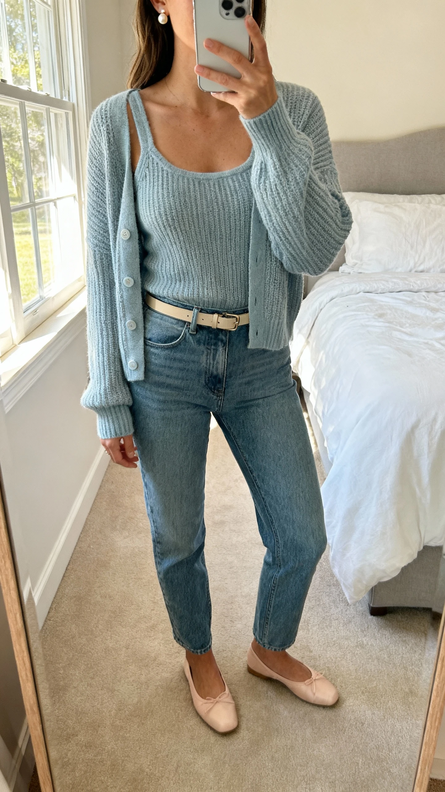 iPhone bedroom mirror selfie of a woman wearing a cardigan set (matching knit tank and cardigan) with straight-leg jeans, ballet flats, thin belt, pearl studs, face not visible, cropped at neck, soft afternoon window light, casual iPhone quality.