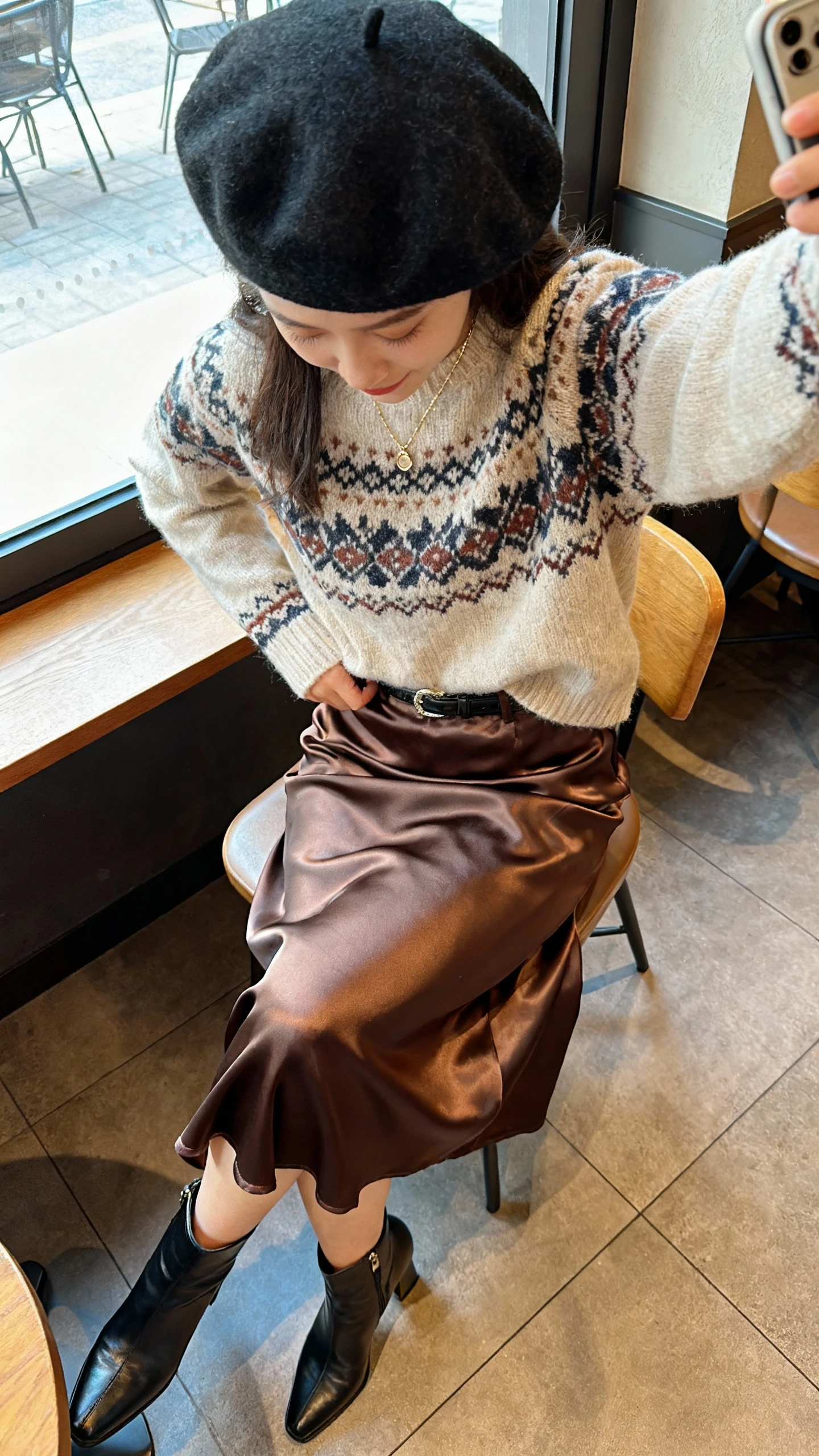 iPhone cafe table selfie (downward angle) of a woman seated wearing a Fair Isle sweater tucked into a satin midi skirt, heeled ankle boots, wool beret, dainty necklace, face not visible, cozy cafe window light, casual iPhone photo.