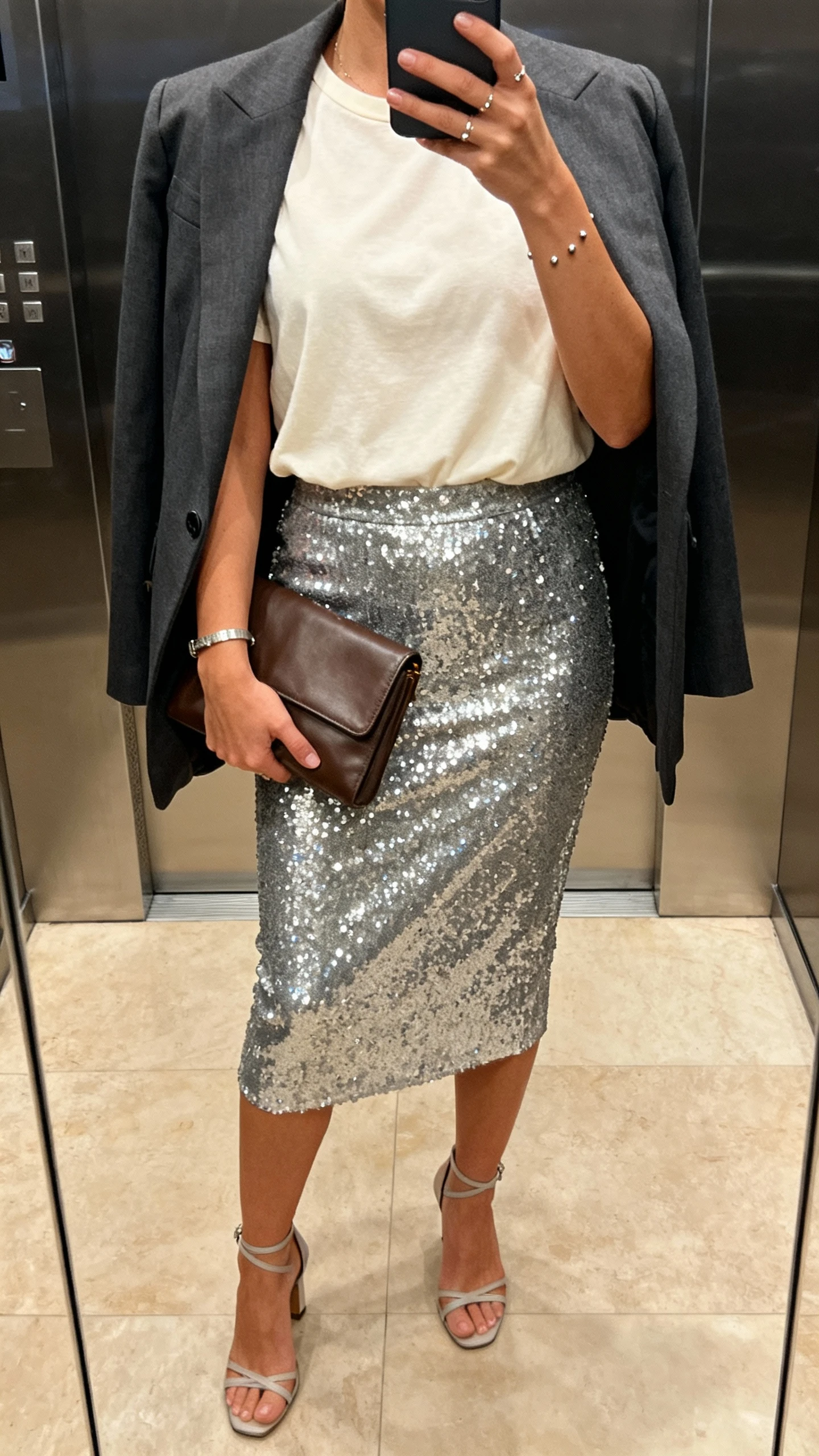 iPhone mirror selfie cropped at neck showing a woman in a silver sequin midi pencil skirt, cream slightly boxy tee, strappy heels, matte leather clutch, delicate studs and simple bracelet, tailored blazer draped over shoulders, face not visible, elevator mirror, evening indoor lighting, natural iPhone photo quality.