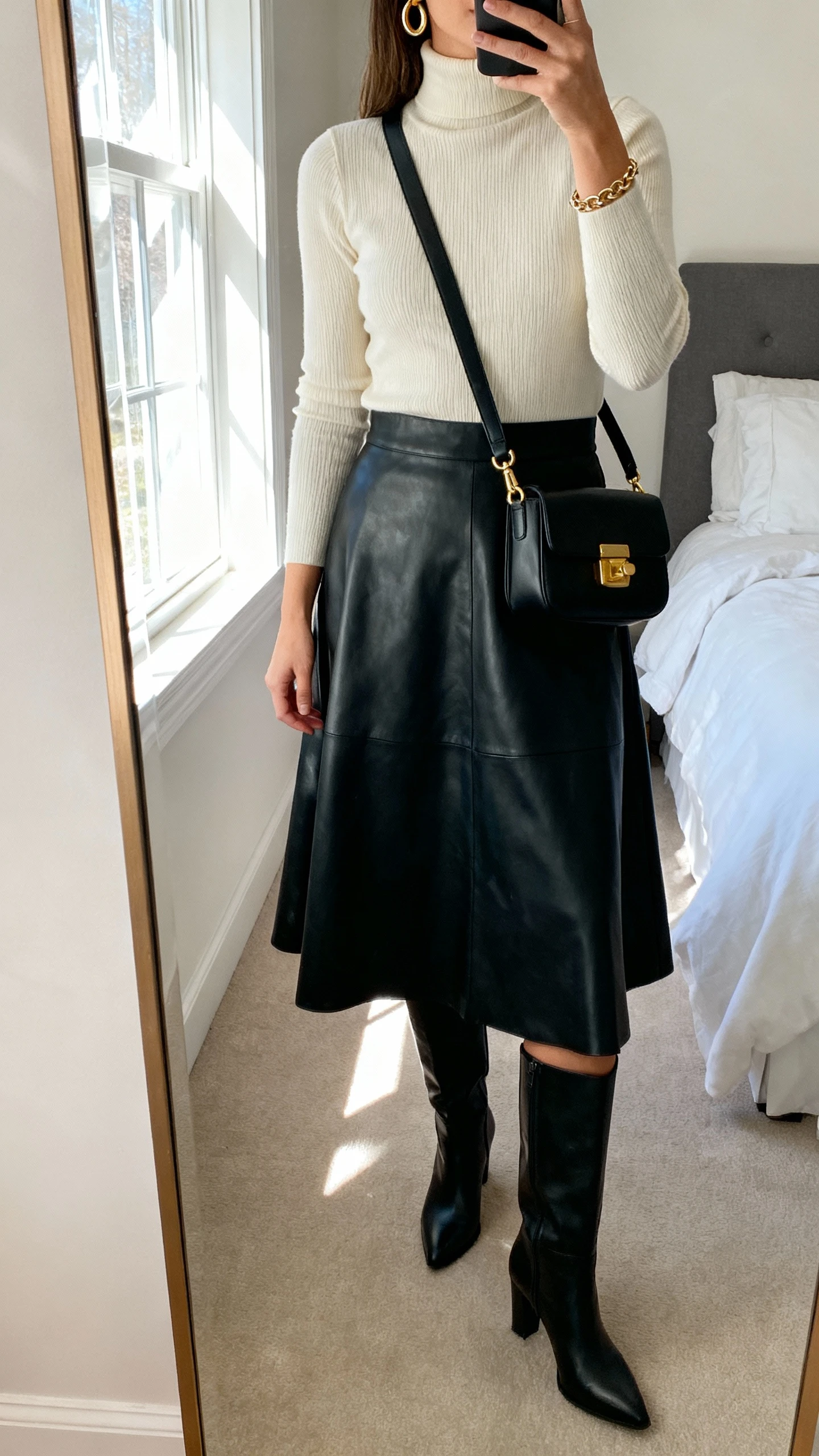 iPhone mirror selfie of a woman in a black leather midi skirt (A-line), cream fine-gauge turtleneck, black knee-high heeled boots, structured crossbody with gold hardware, gold hoop earrings and delicate chain bracelet; face not visible, cropped at neck, bedroom mirror with window light, natural iPhone photo quality.