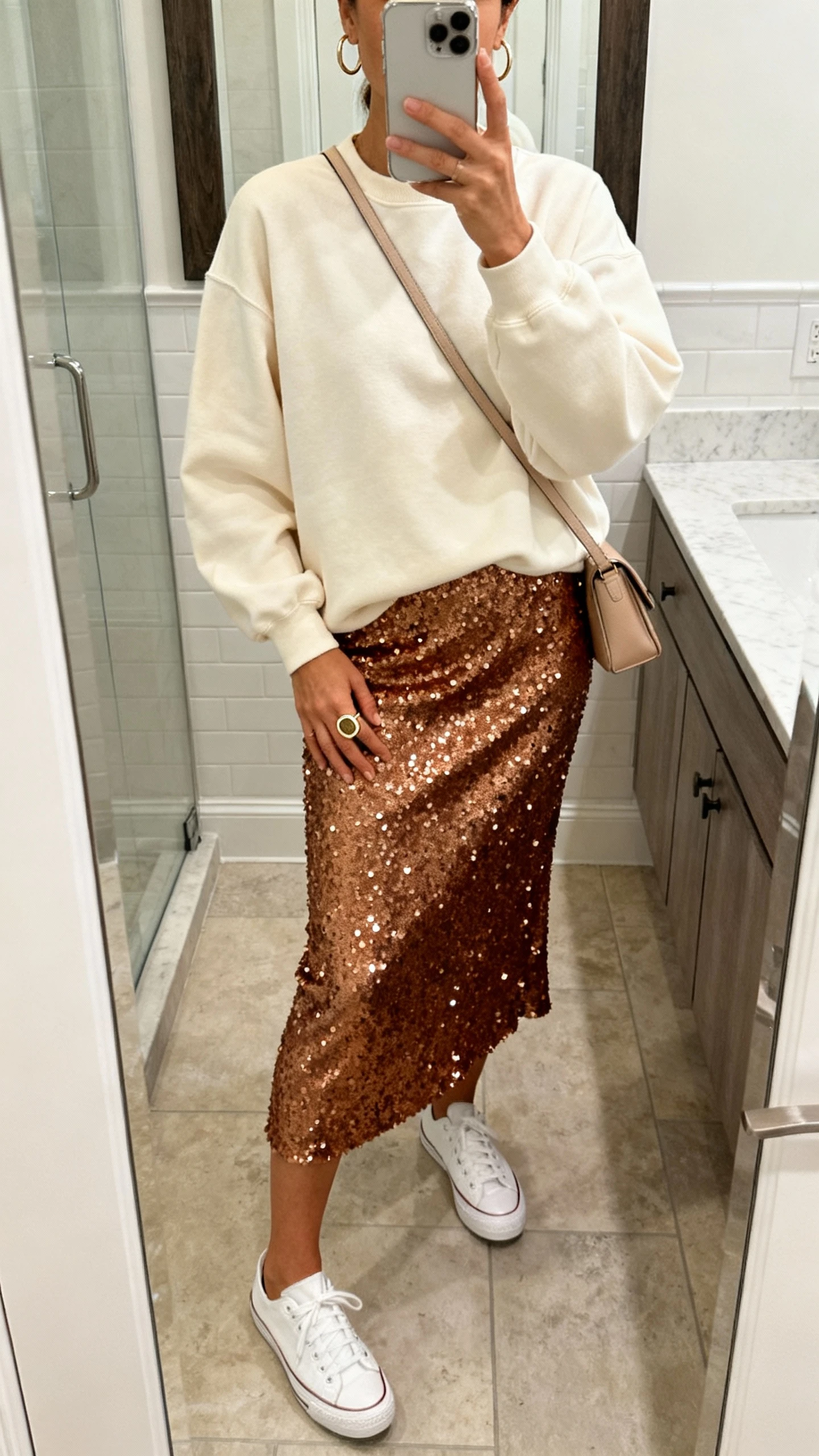 iPhone mirror selfie of a woman in a bronze sequin midi skirt, relaxed cream crewneck sweatshirt, sleek white sneakers, minimal crossbody, simple hoops and one statement ring; face not visible, bathroom mirror, soft indoor lighting, casual iPhone photo quality.