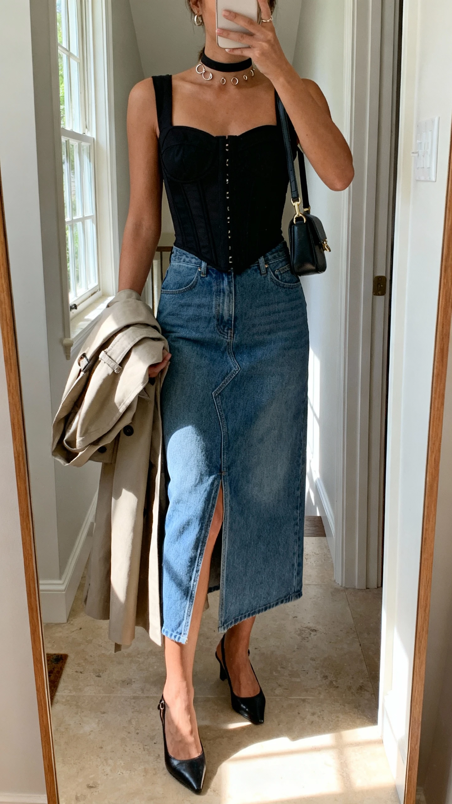 iPhone mirror selfie of a woman in a denim midi skirt with front slit, black corset top, slingback heels, mini shoulder bag, choker with small hoops, lightweight trench draped over arm, face not visible, entryway mirror, daylight from window, natural iPhone photo.