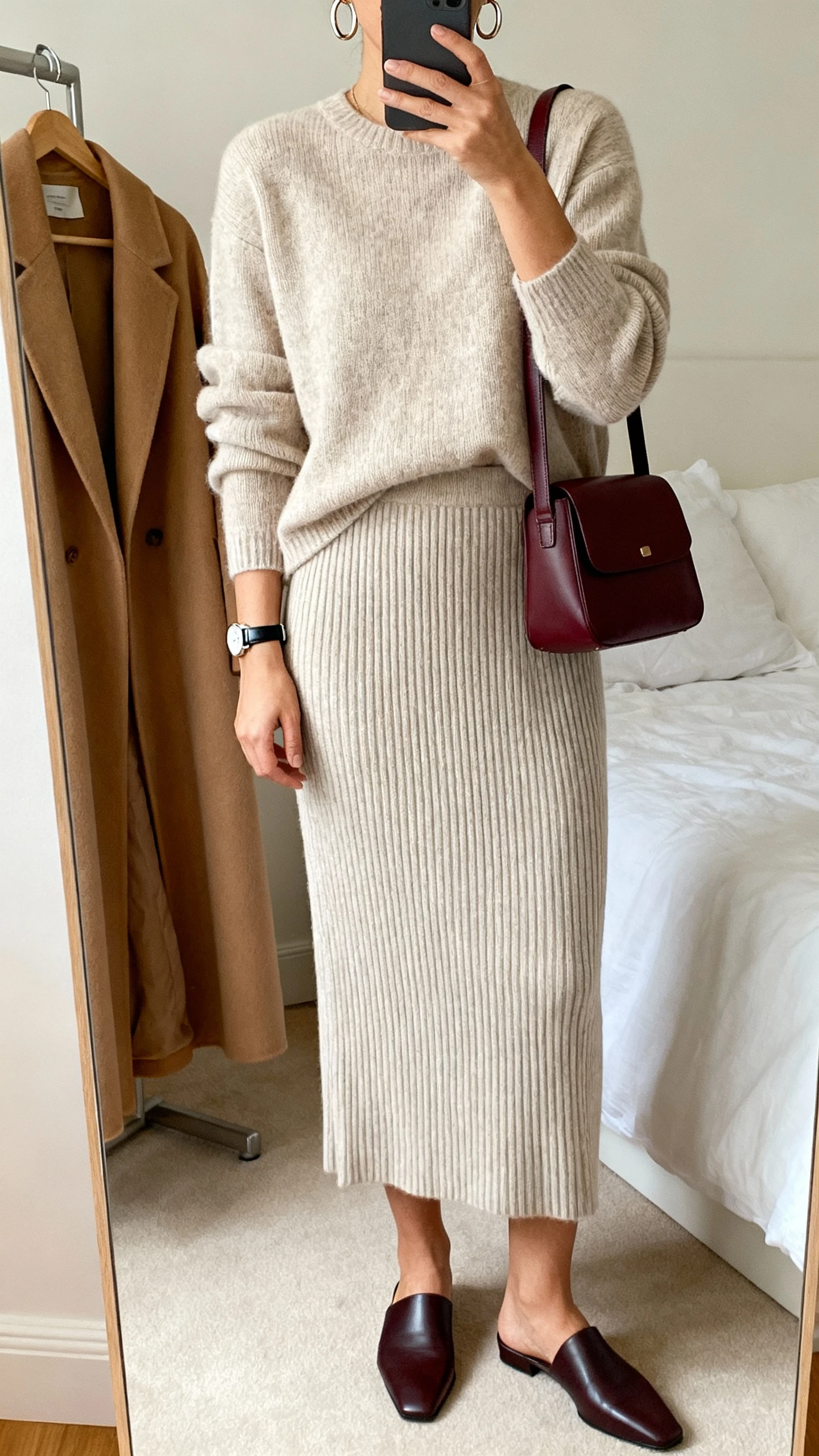 iPhone mirror selfie of a woman in a ribbed knit column midi skirt with matching knit long-sleeve top, sleek mules, oxblood shoulder bag, hoops and a sleek watch, camel longline coat on hanger nearby, face not visible, bedroom mirror, soft morning light, natural iPhone photo.