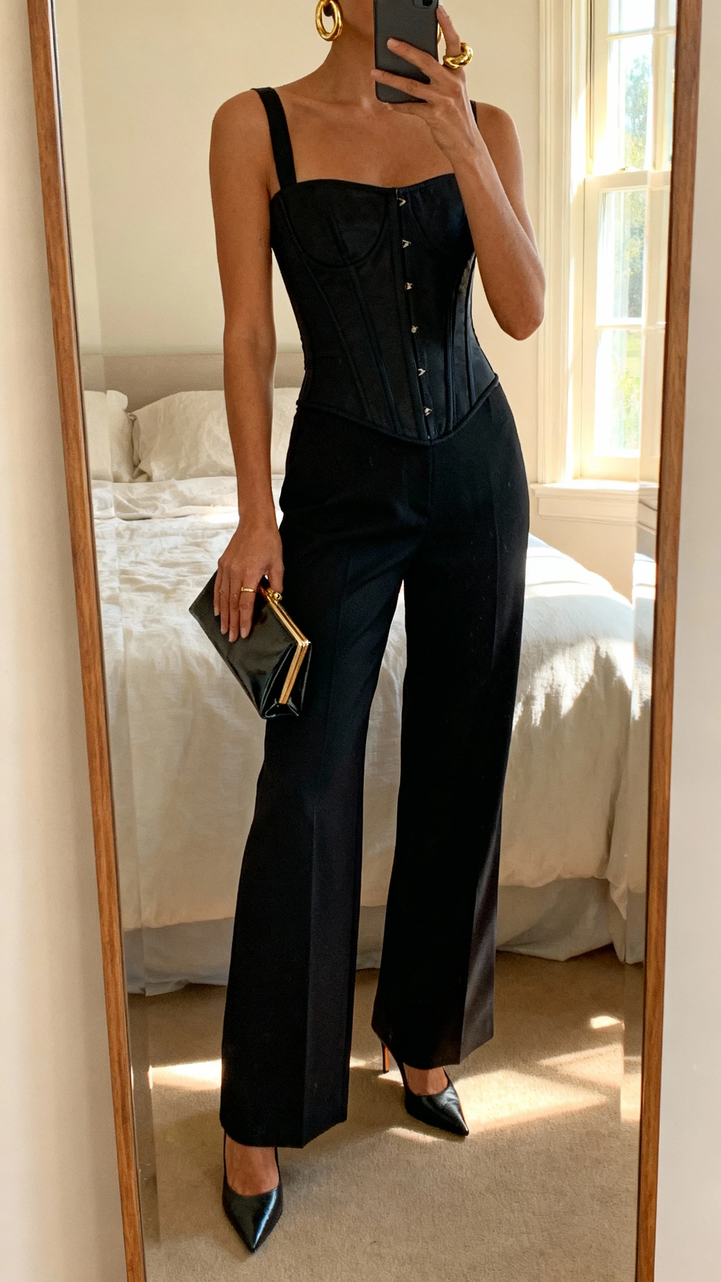 iPhone mirror selfie of a woman in a structured black corset top, high-waisted wide-leg trousers, pointed-toe heels, chunky gold hoops, and a sleek clutch, face not visible, cropped at neck, bedroom mirror with soft window light, natural iPhone photo quality