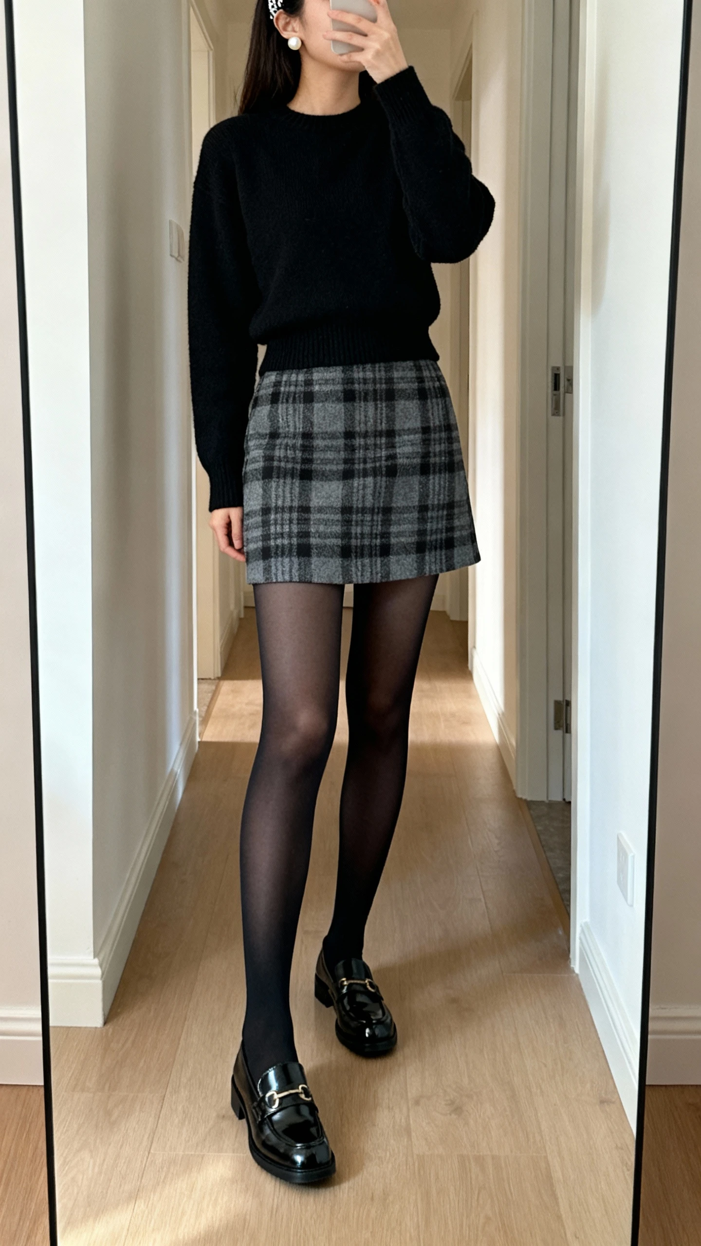 iPhone mirror selfie of a woman wearing a gray/black plaid mini skirt, black opaque tights, fitted black crewneck sweater, patent loafers, headband and pearl studs; face not visible, cropped at shoulders, hallway full-length mirror, soft morning light, natural iPhone quality.