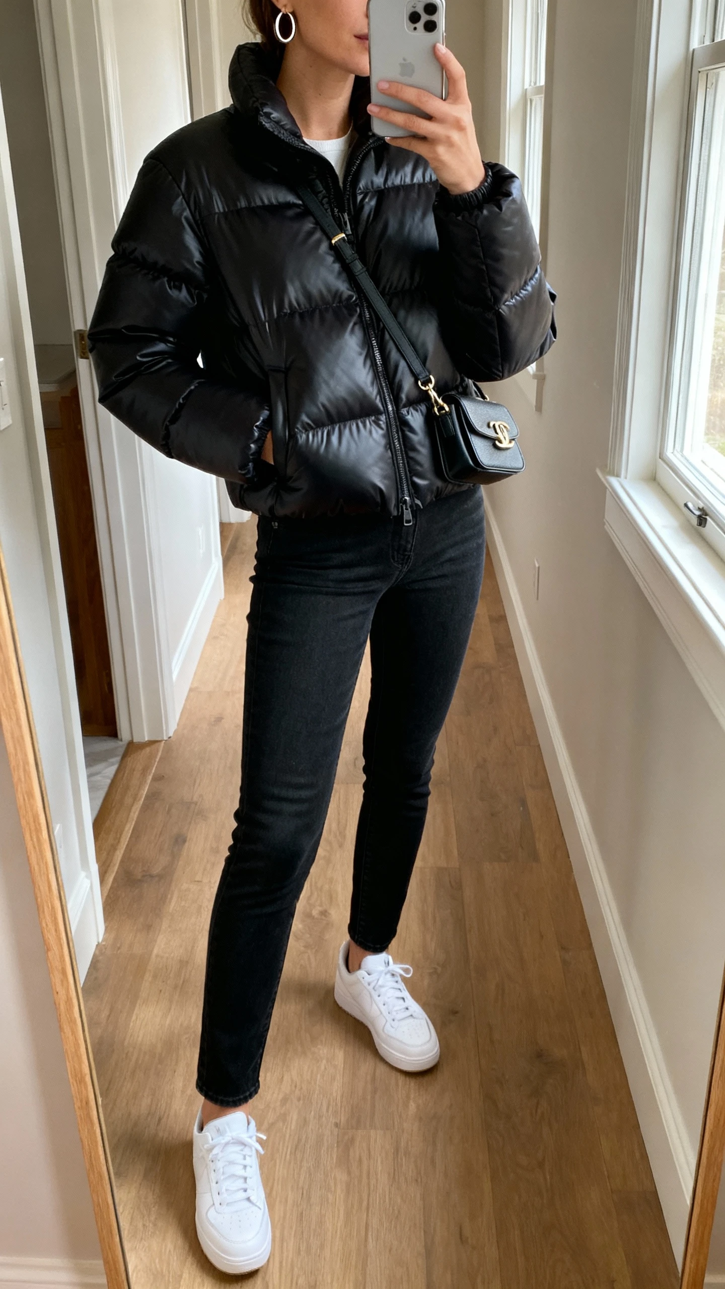 iPhone mirror selfie of a woman wearing a sleek matte-black city-walk puffer jacket that looks designer, slim black jeans, white sneakers, small crossbody bag, simple hoop earrings, face not visible, cropped at neck, hallway mirror with morning window light, natural iPhone photo quality