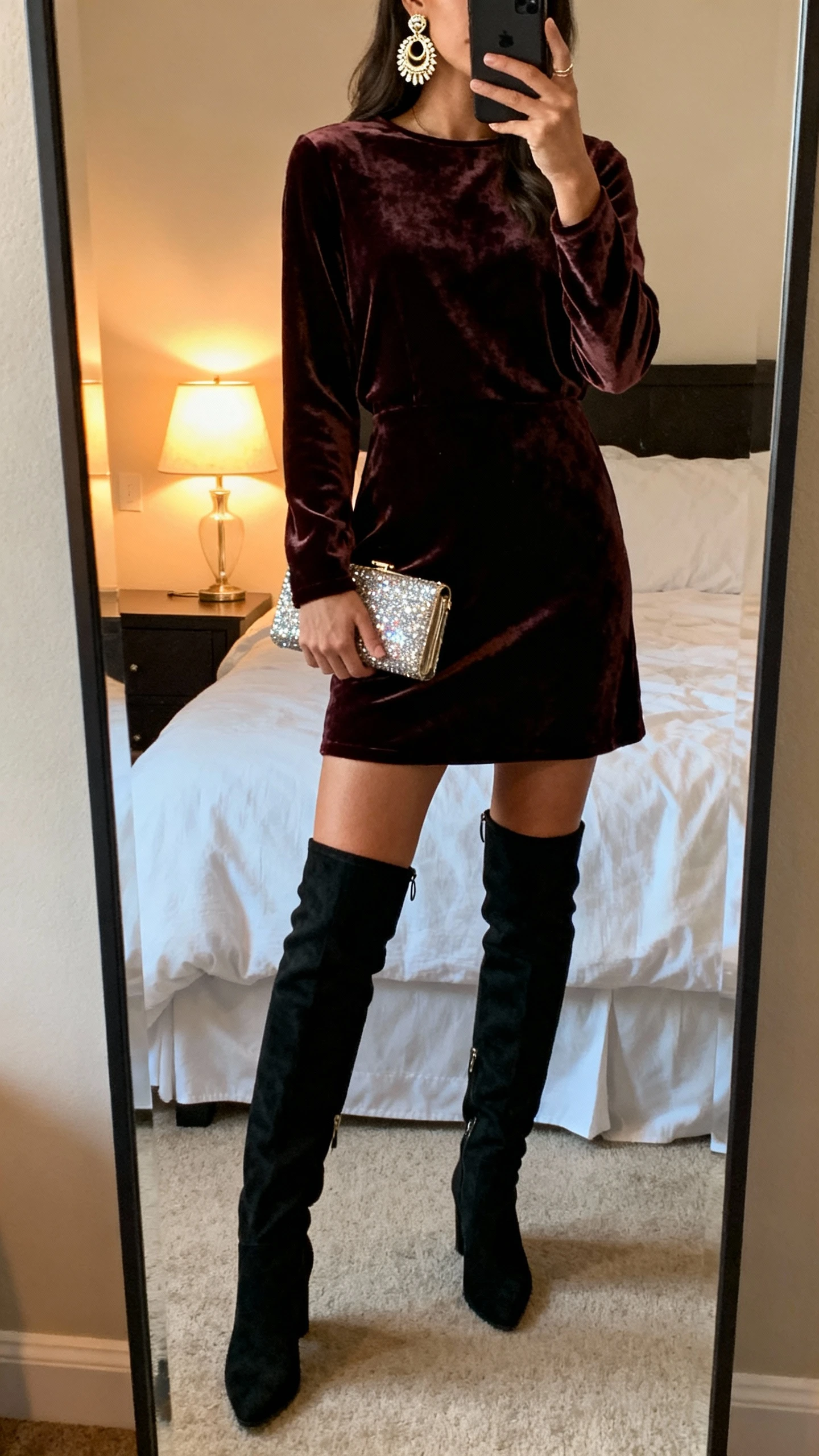 iPhone mirror selfie of a woman wearing a velvet mini dress with over-the-knee boots, statement earrings, and a small sparkly clutch, face not visible, bedroom full-length mirror, warm lamp light, natural iPhone photo quality