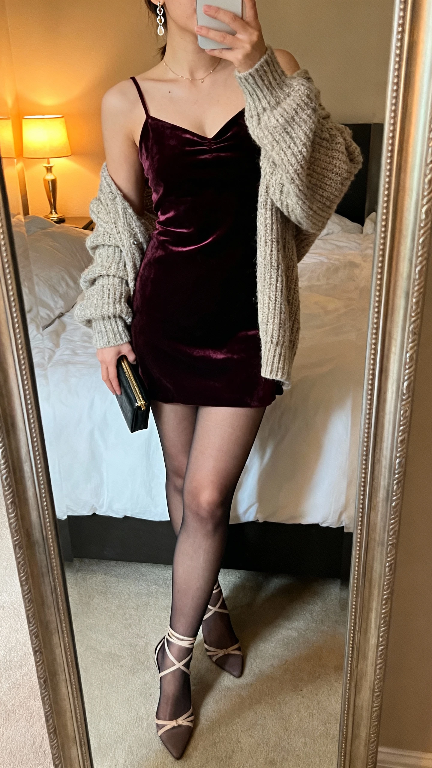 iPhone mirror selfie of a woman wearing a velvet slip dress layered under a chunky sweater, sheer tights, strappy heels, delicate earrings, small clutch in hand, face not visible, cropped at neck, bedroom mirror with warm lamp light, natural iPhone photo quality.