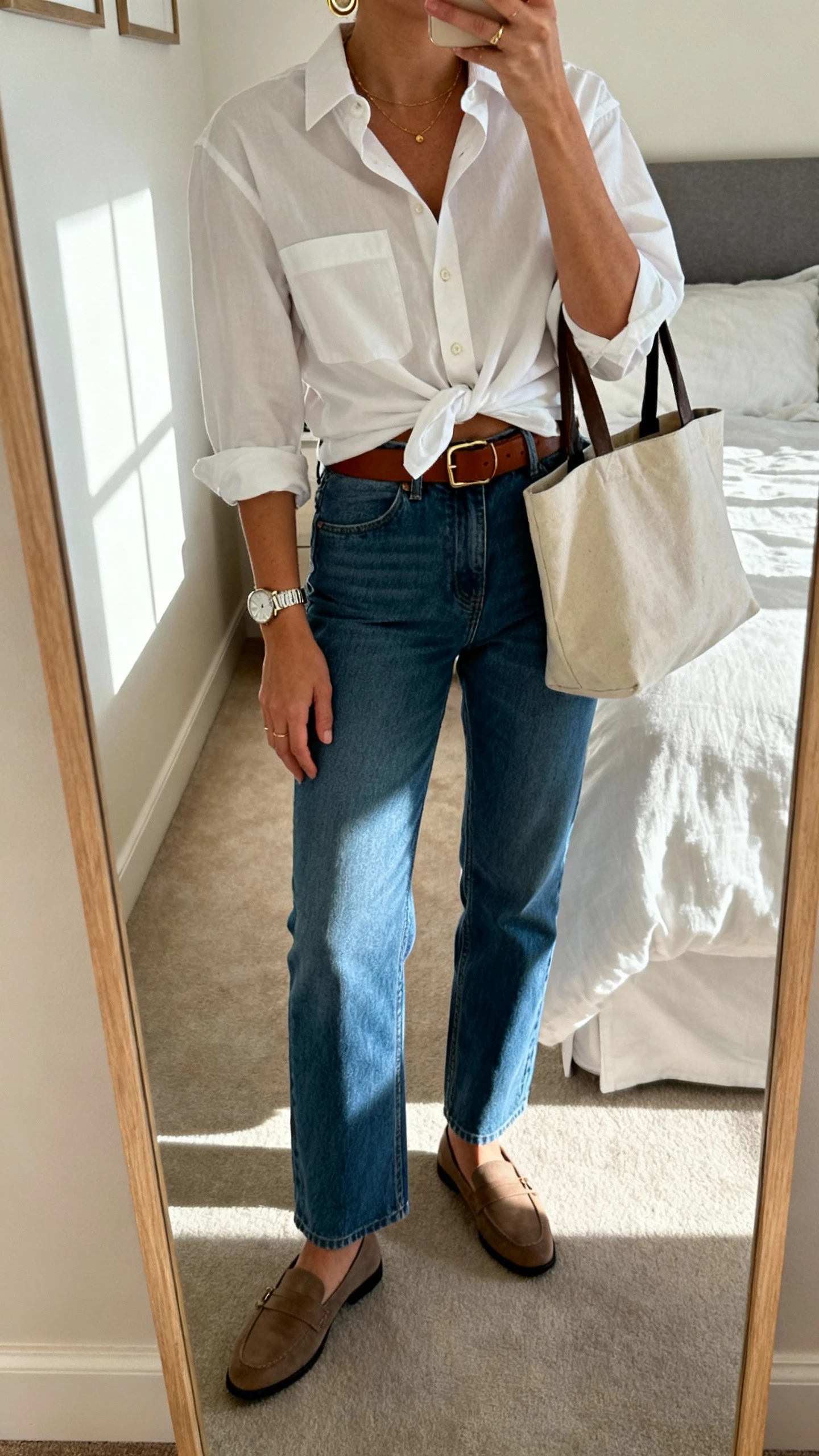 iPhone mirror selfie of a woman wearing straight-leg blue jeans, crisp white button-down half-tucked, brown belt, loafers, delicate gold hoops and watch, tote bag on arm, face not visible, cropped at neck, bedroom mirror with morning window light, natural iPhone photo quality.