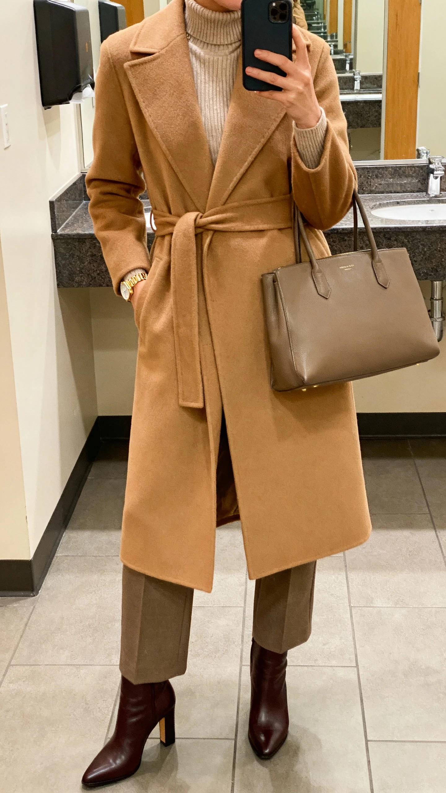iPhone office bathroom mirror selfie of a woman in a belted camel wool coat over a fine knit turtleneck, tailored trousers, heeled ankle boots, structured tote, minimal gold watch, face not visible, cropped at shoulders, warm indoor lighting, natural iPhone photo quality
