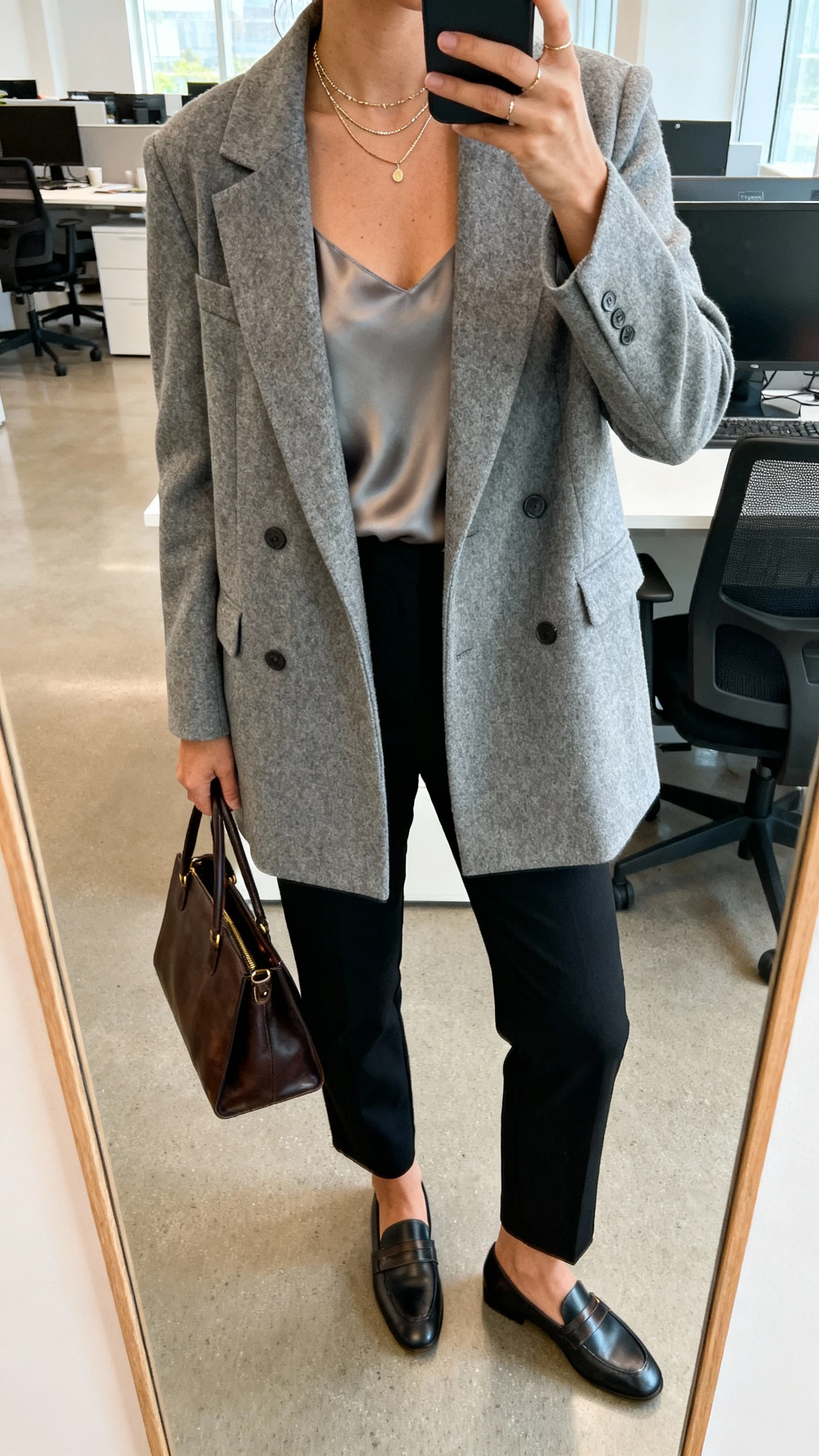 iPhone office mirror selfie of a woman wearing a chic blazer-coat hybrid in heather gray, silk camisole, straight-leg black trousers, loafers, leather tote, delicate layered necklaces, face not visible, cropped at neck, bright morning office light, natural iPhone photo quality