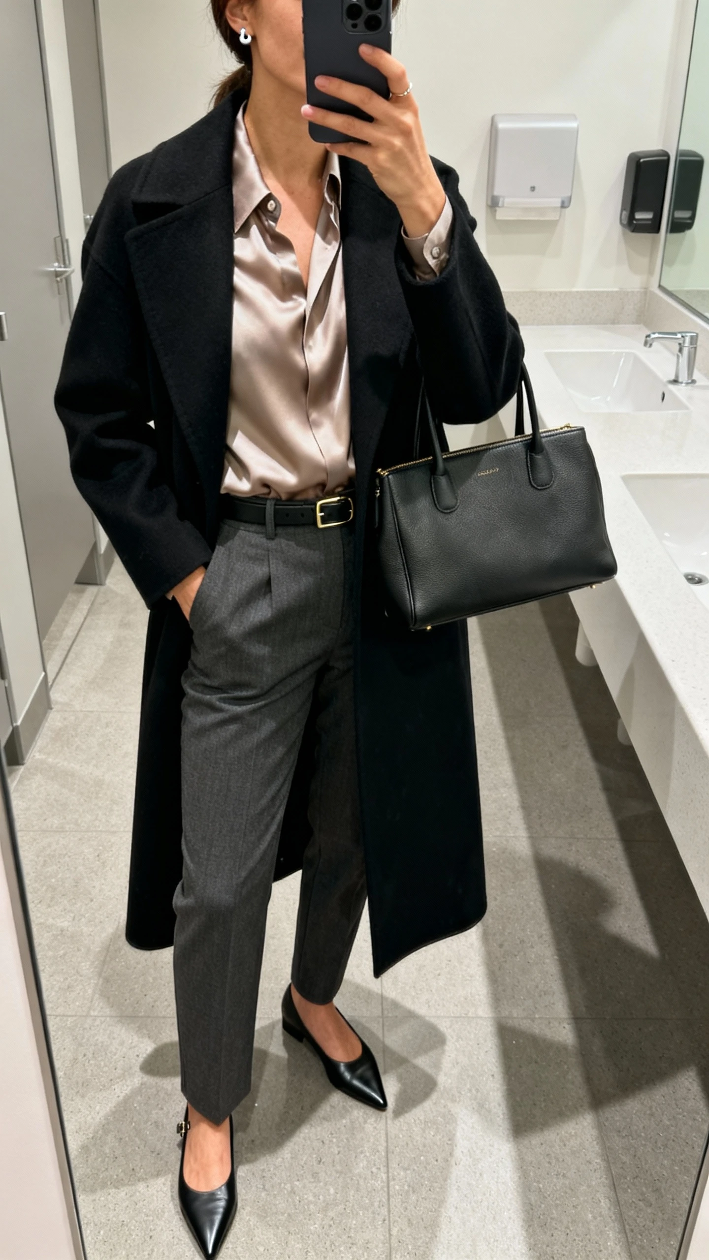 iPhone office restroom mirror selfie of a woman in tailored trousers, a silk blouse, and a longline coat, pointed-toe loafers, slim belt, minimal stud earrings, structured tote, face not visible, natural overhead lighting, casual iPhone aesthetic.