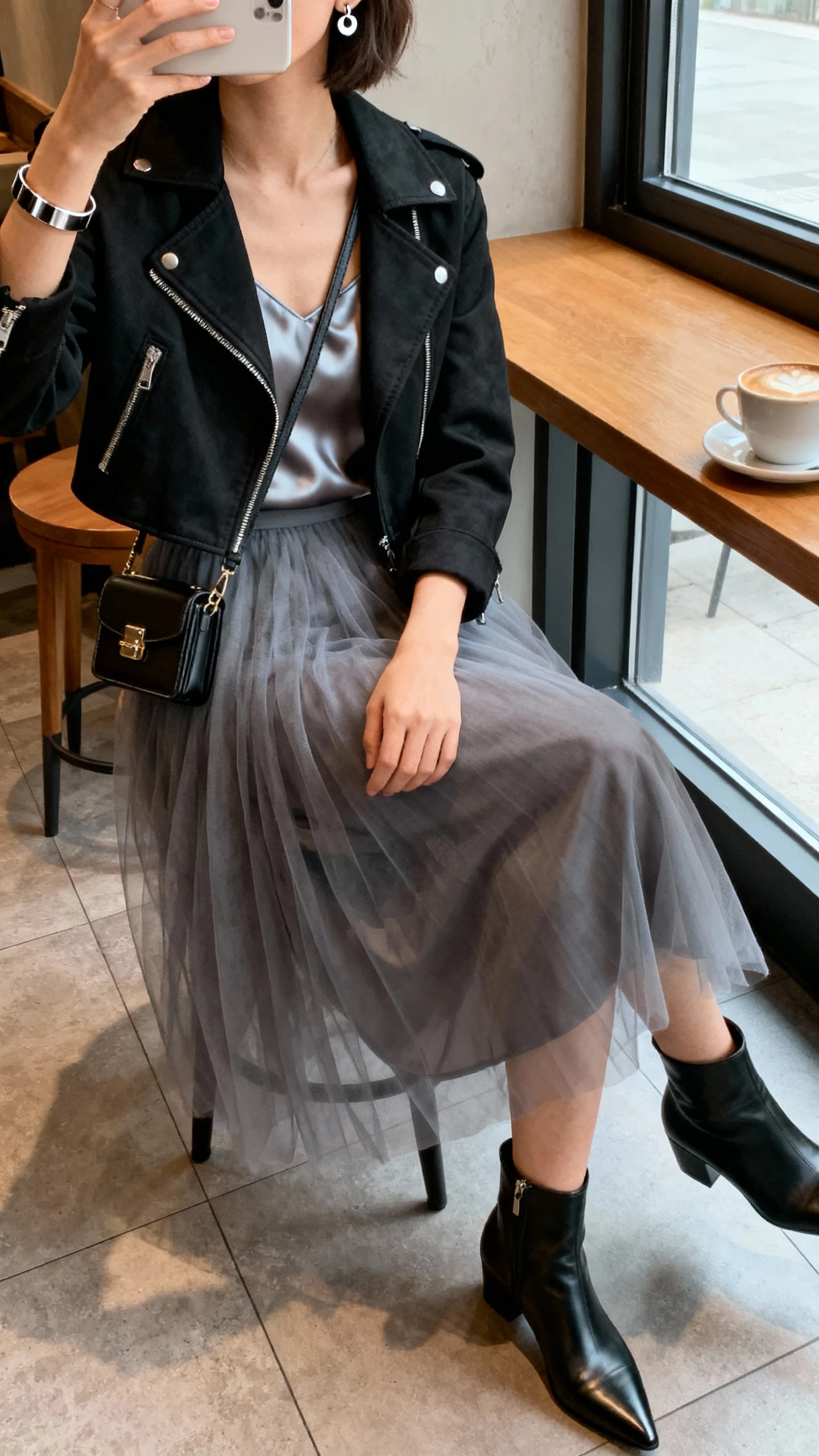 iPhone seated cafe selfie cropped at shoulders showing a woman wearing a smoke gray tulle midi skirt, satin camisole, cropped black moto jacket, pointed ankle boots, compact crossbody, stud earrings and cuff bracelet, face not visible, window light at coffee shop, natural iPhone quality.