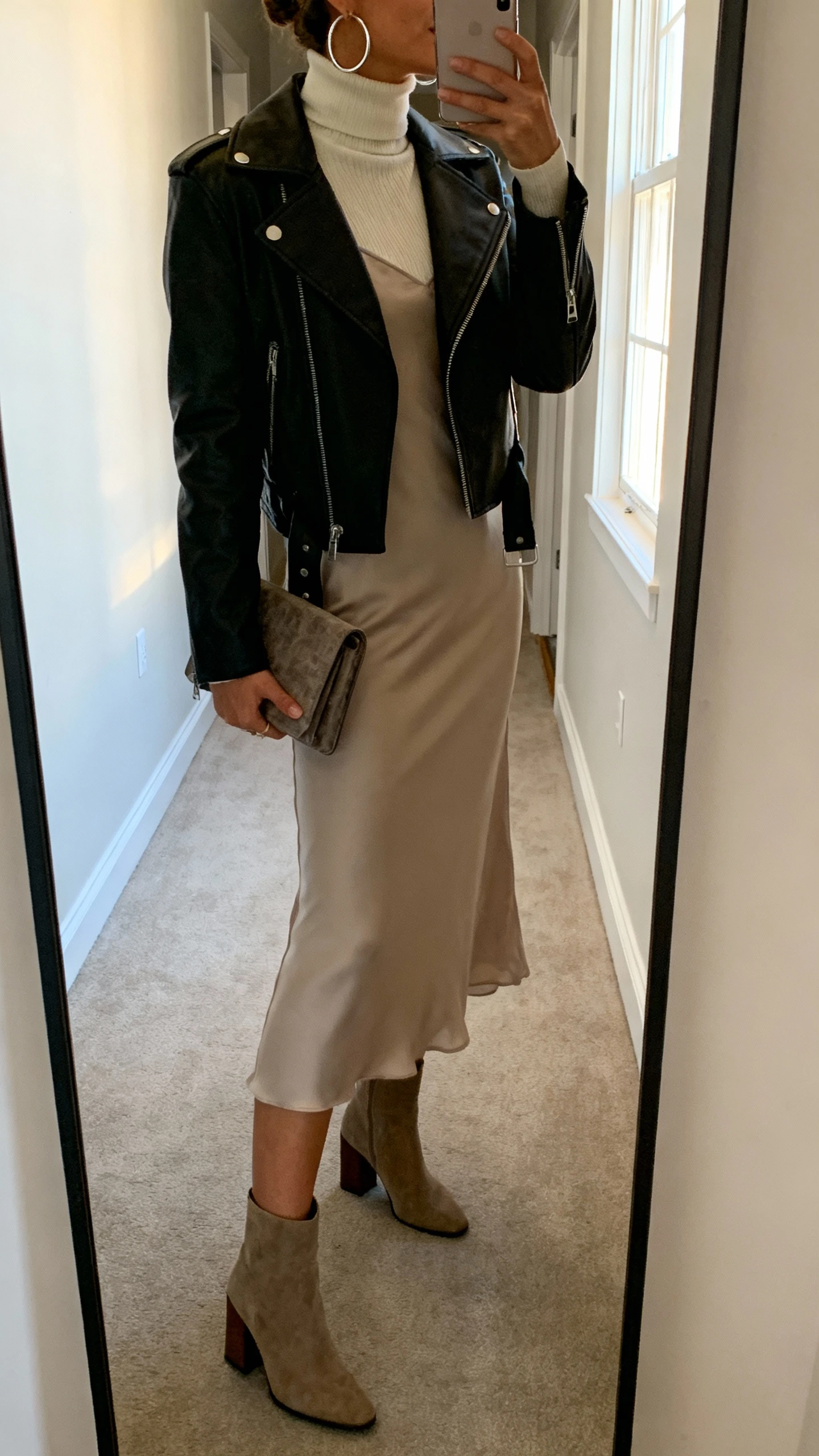 iPhone side-profile mirror selfie of a woman layering a turtleneck under a slip dress with a moto jacket on top, block-heel boots, hoop earrings, clutch, face not visible, apartment hallway mirror with window light, casual iPhone aesthetic.