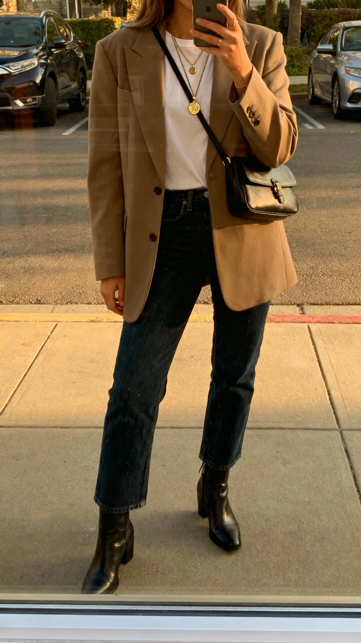 iPhone street-mirror window selfie of a woman in a statement blazer, dark denim, and sleek ankle boots, simple tee underneath, layered pendant necklace, structured crossbody, face not visible, late afternoon daylight, natural iPhone quality.