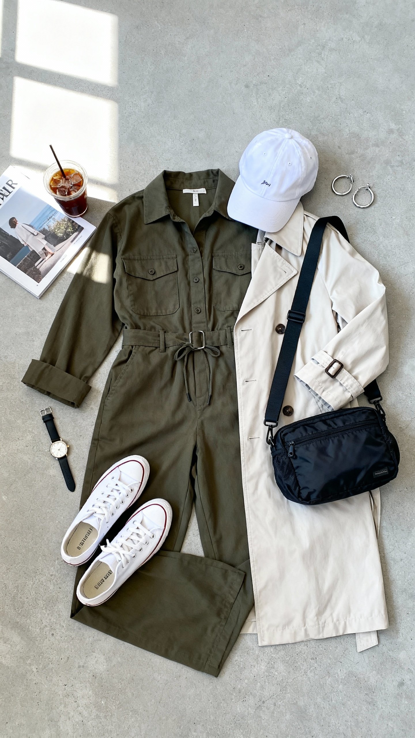 Flat lay overhead view of weekend jumpsuit with slick layering: utility jumpsuit, lightweight trench coat, white sneakers, baseball cap, crossbody nylon bag, silver hoop earrings and watch on light gray concrete surface, magazine and iced coffee as props, bright natural window light, organized composition, no person visible, iPhone photo