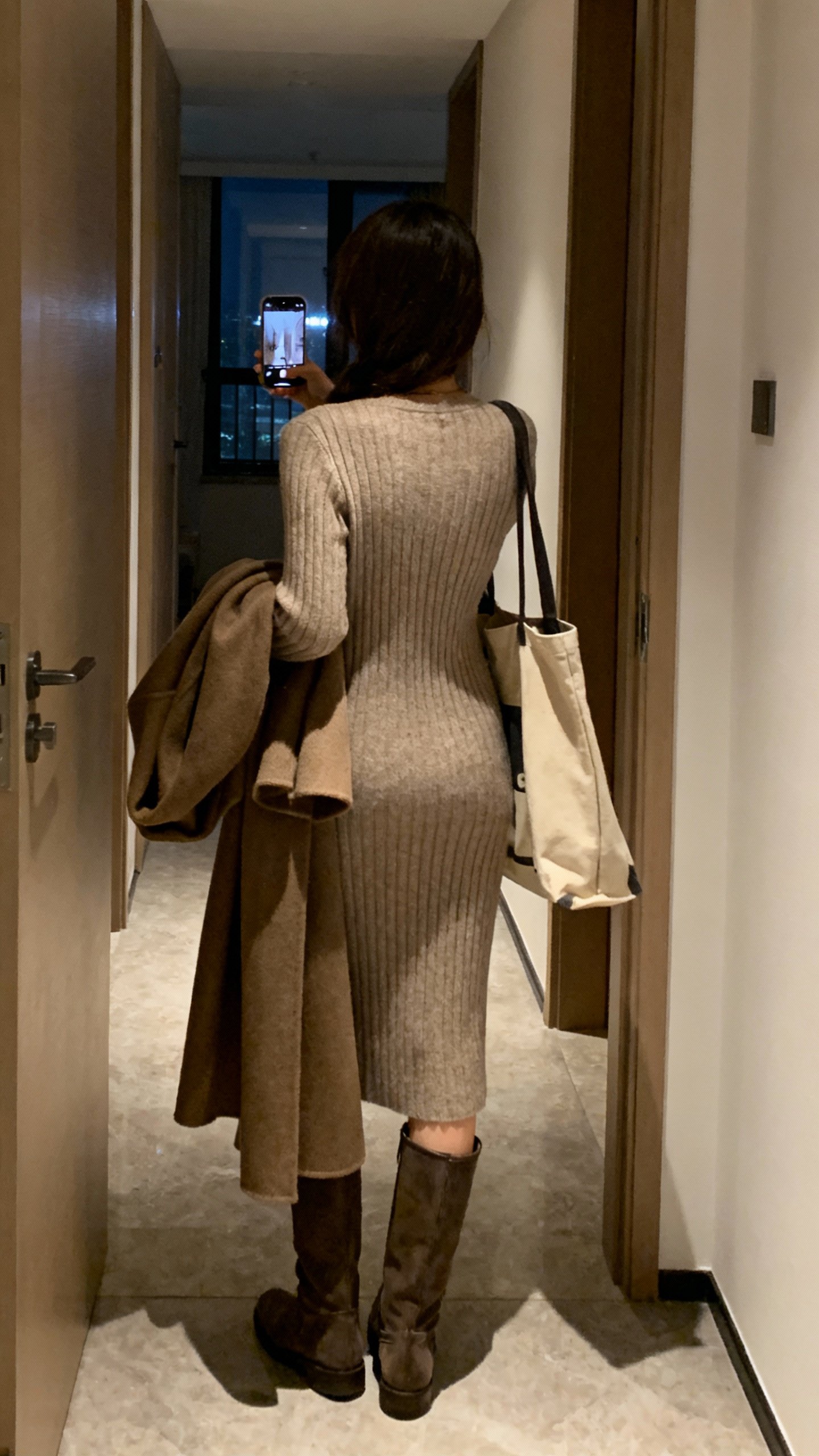 iPhone back-view hallway selfie showing a cozy ribbed knit dress with knee-high boots and a long coat draped over arm, tote bag, face not visible, indoor evening lighting, casual iPhone quality.