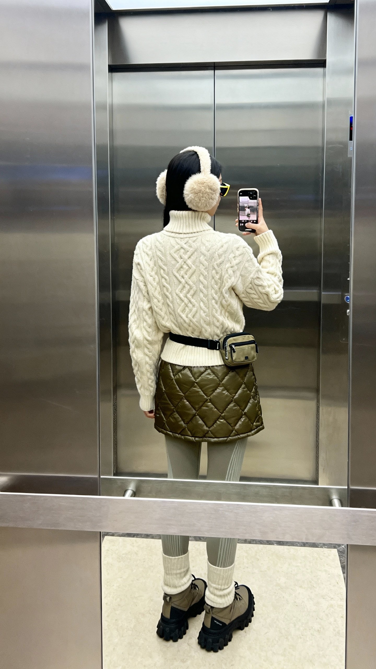 iPhone back-view selfie in elevator mirror: cream cable-knit turtleneck, quilted nylon mini skirt over thermal leggings, après-ski faux-fur ear muffs, lug-sole boots, sporty sunglasses, mini belt bag; cool daylight, face not visible, natural iPhone photo.