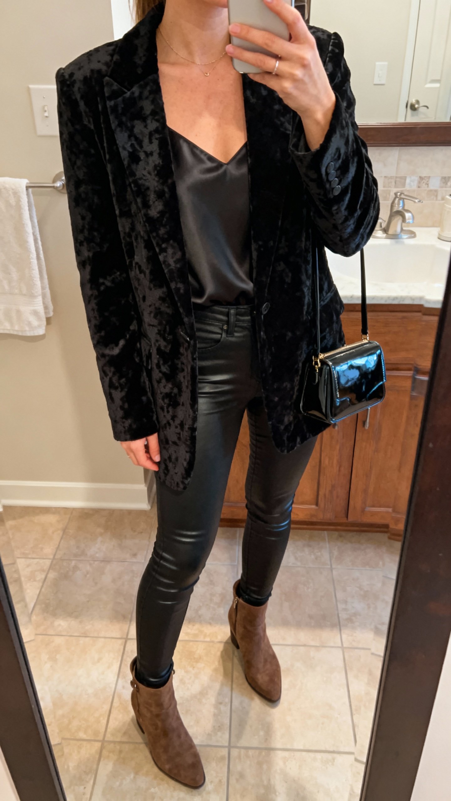iPhone bathroom mirror selfie in all-black: textured velvet blazer, silk camisole, coated skinny jeans, suede ankle boots, patent mini bag, layered black textures, face not visible, cropped at neck, warm indoor lighting, natural iPhone photo quality.
