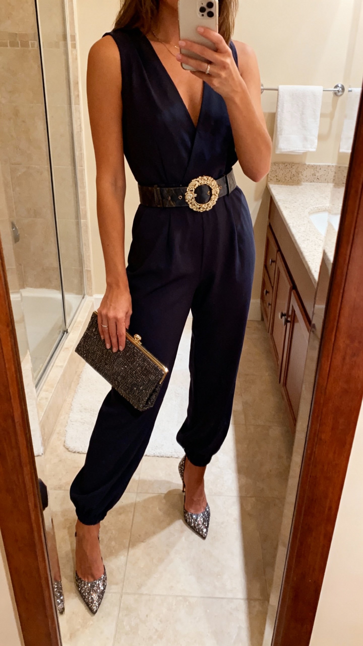 iPhone bathroom mirror selfie of an elevated jumpsuit cinched with a statement belt and sparkly heels, clutch in hand, face not visible, warm indoor lighting, natural iPhone quality.