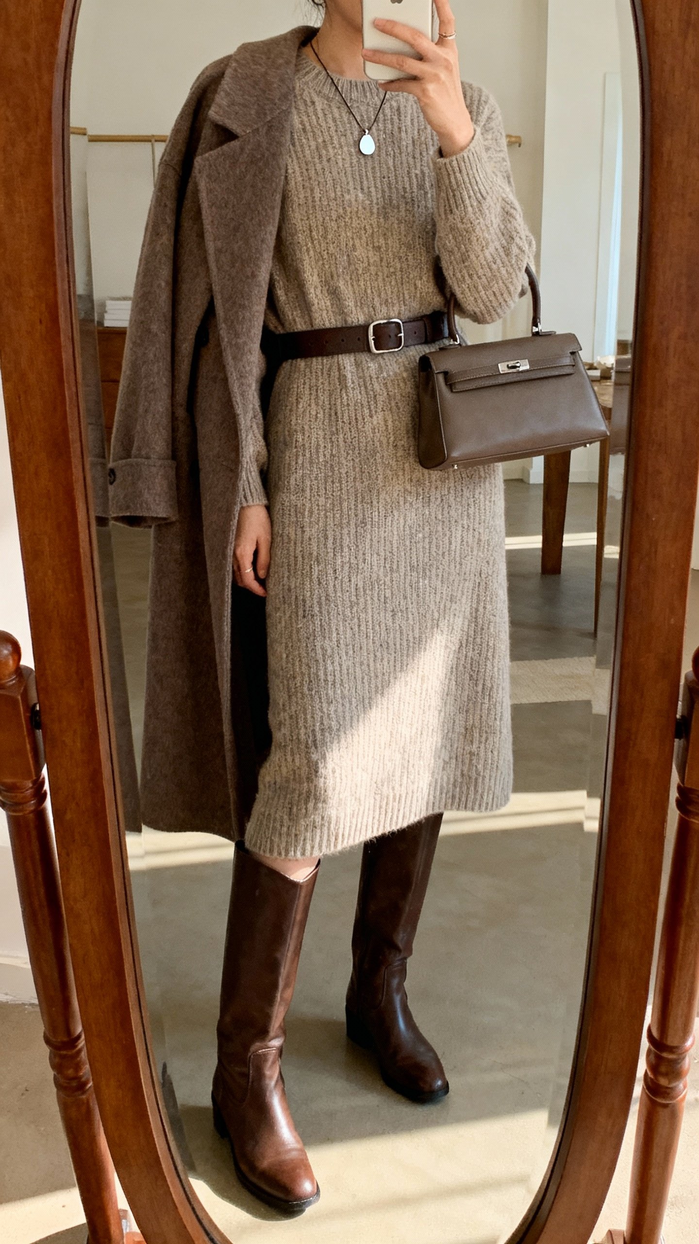 iPhone boutique mirror selfie featuring a knit midi dress, tall leather boots, waist-cinching belt, long wool coat draped over shoulders, simple pendant necklace, small top-handle bag; warm indoor lighting, face not visible, casual iPhone quality.