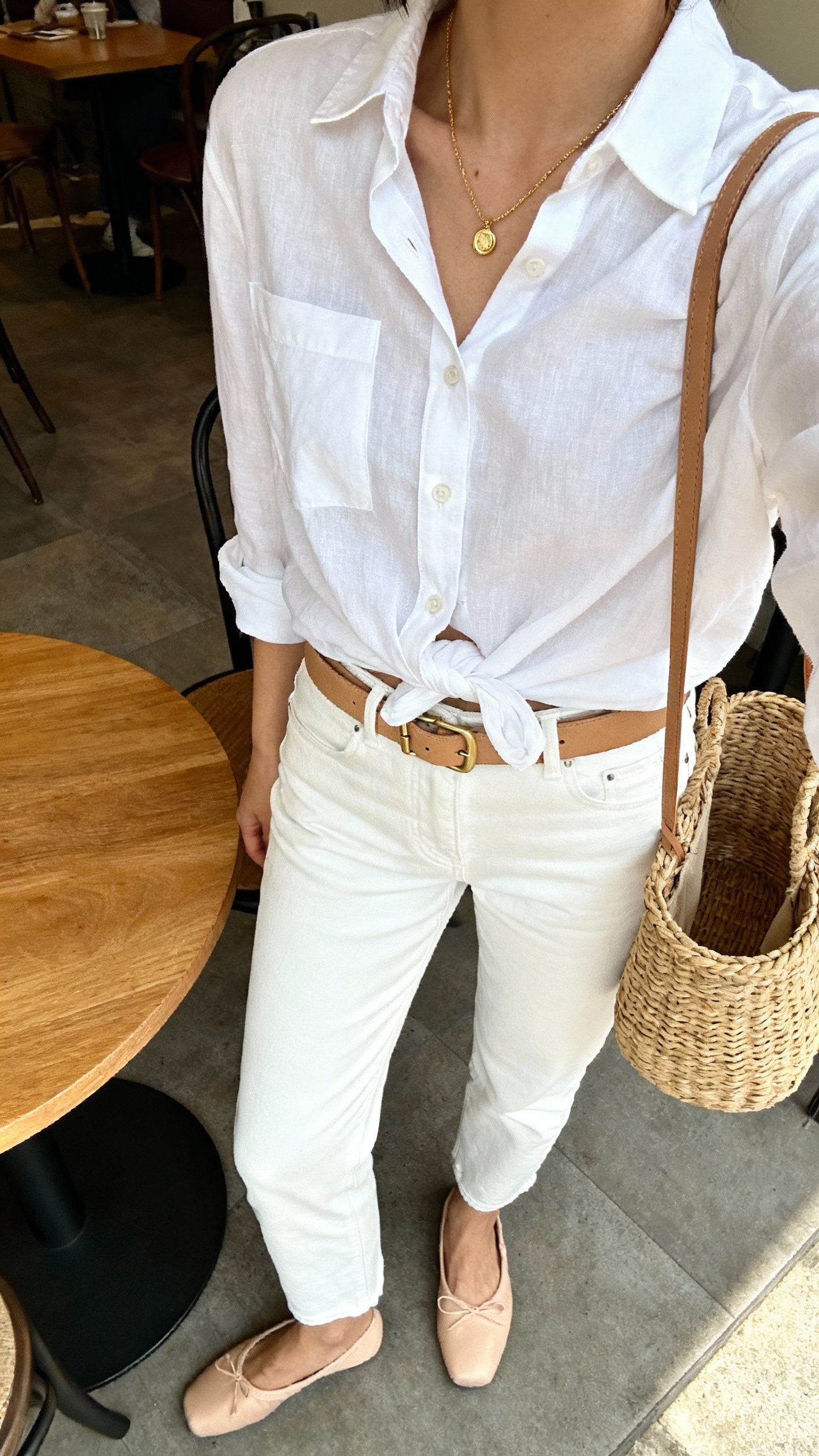 iPhone café table selfie cropped at neck showing a woman in crisp white jeans, relaxed white button-up half-tucked, tan belt, ballet flats, dainty gold necklace, and a wicker basket bag, face not visible, natural daylight, casual iPhone photo.
