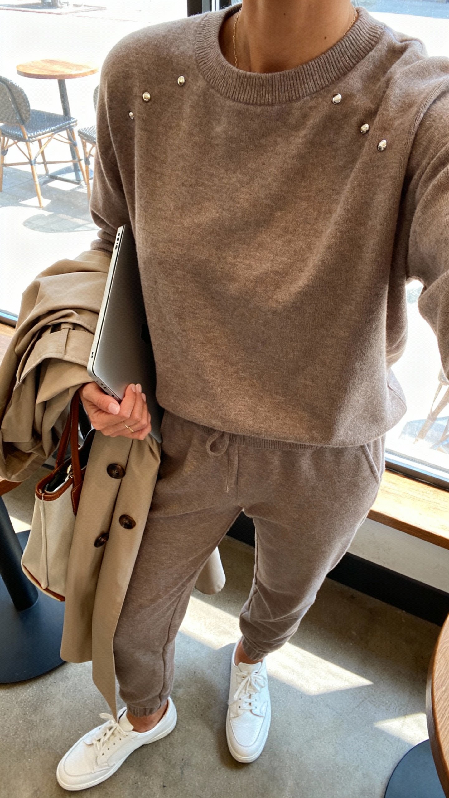 iPhone cafe table selfie cropped at shoulders showing an elevated matching knit sweatsuit (fine-gauge crewneck + tapered joggers) in taupe, white leather sneakers, trench coat over arm, tote with laptop peeking, subtle studs, face not visible, bright cafe window light, natural iPhone quality.