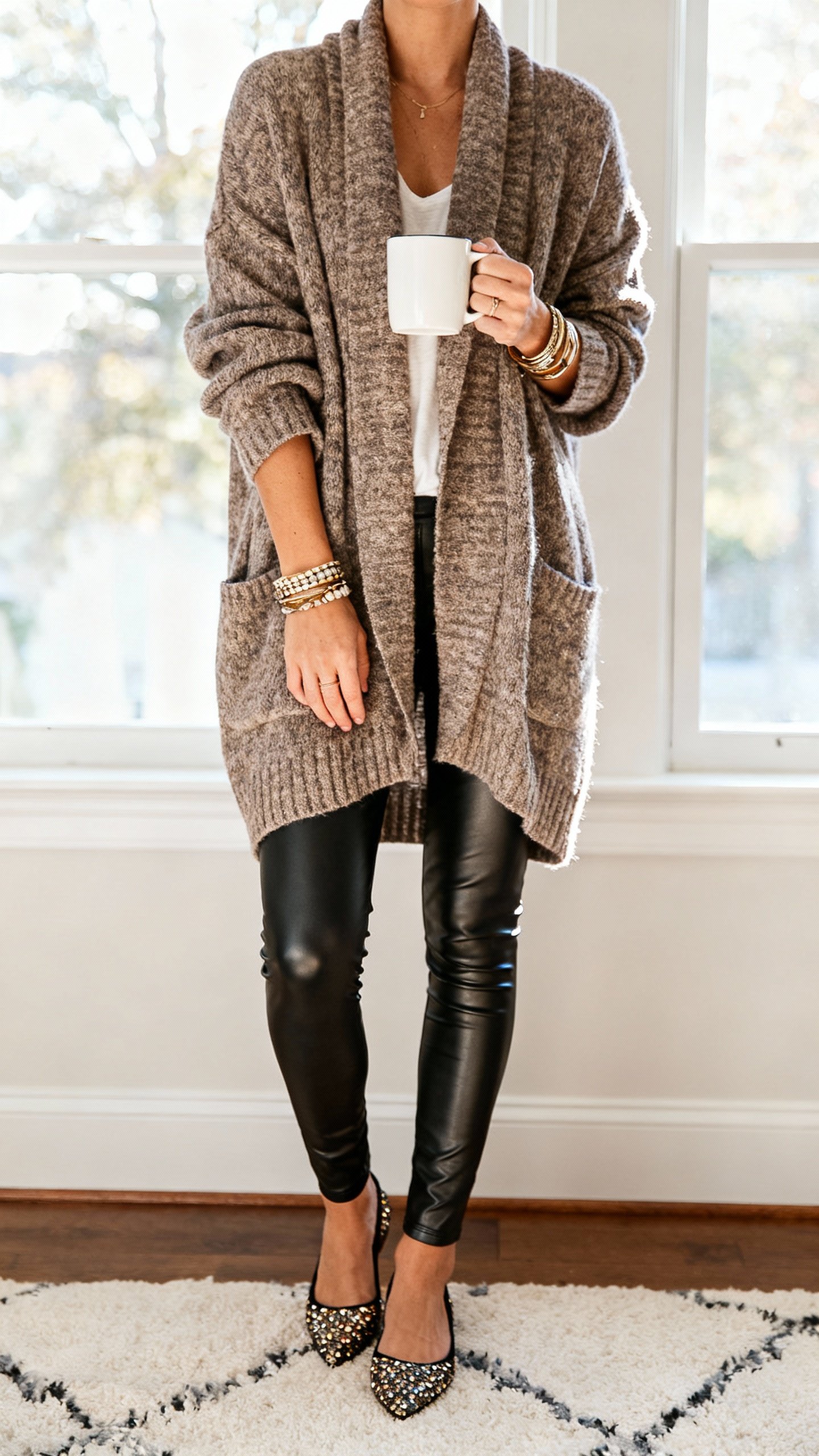 iPhone couch selfie cropped at neck showing faux-leather leggings with an oversized cardigan, glam flats, layered bracelets, coffee mug in hand, face not visible, soft afternoon window light, natural iPhone quality.