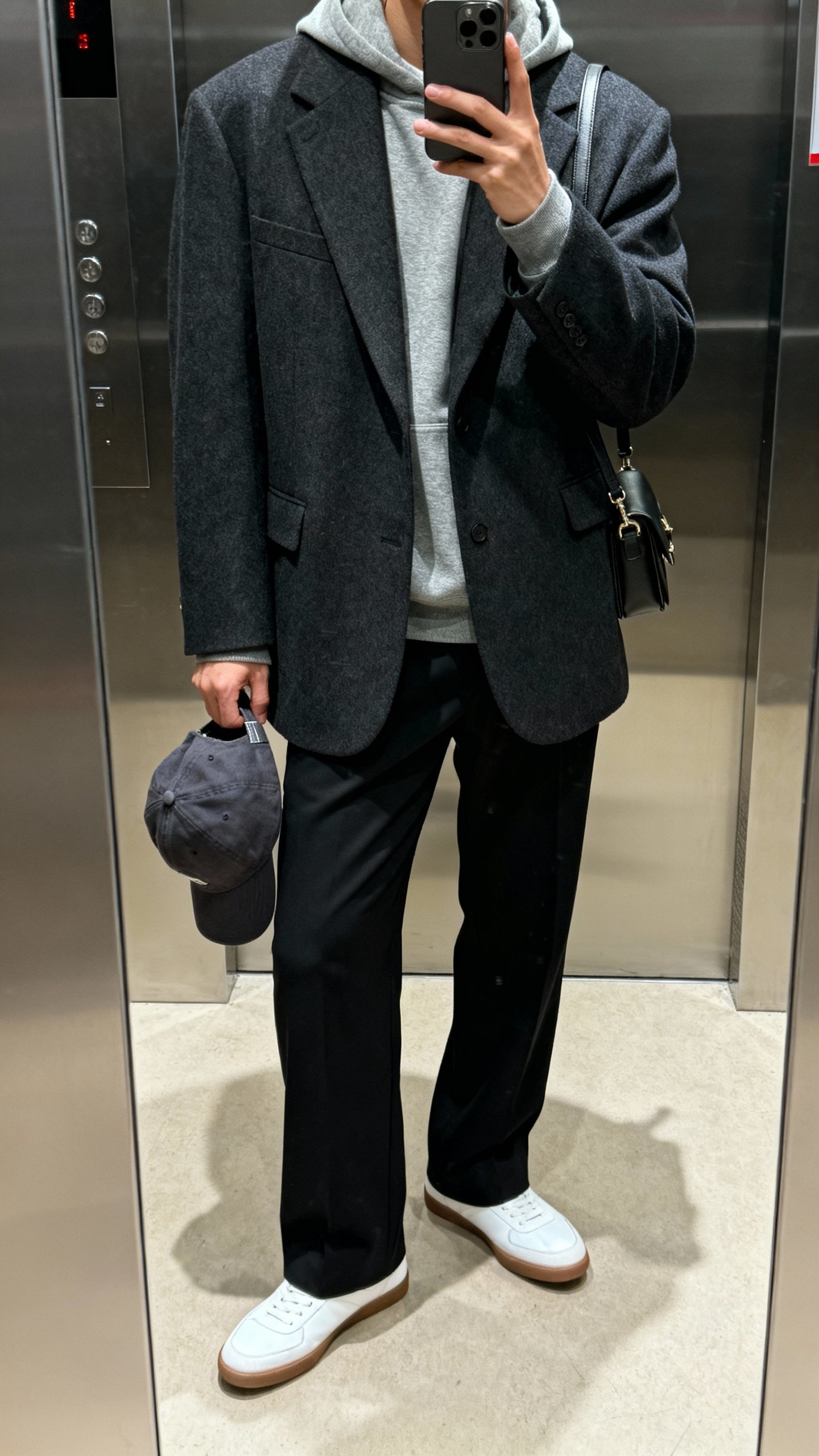 iPhone elevator mirror selfie of an oversized charcoal blazer layered over a gray hoodie with tailored black trousers, white low-profile sneakers, baseball cap in hand, small shoulder bag, face not visible, indoor lighting, casual iPhone quality.