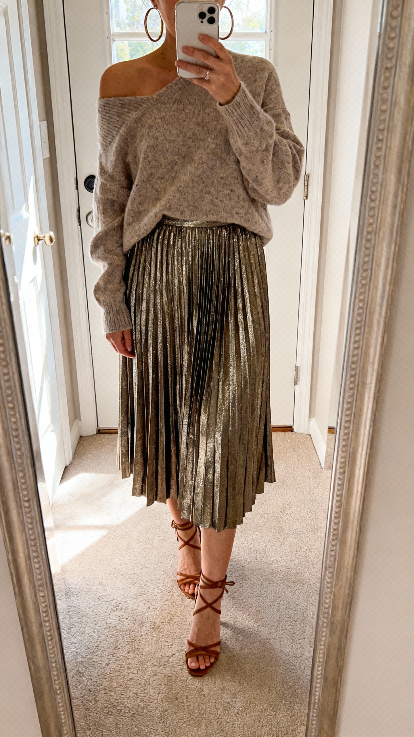 iPhone full-length mirror selfie showing a metallic pleated midi skirt with an off-shoulder sweater, strappy heels, hoop earrings, face not visible, entryway mirror with daylight from door, natural iPhone photo.