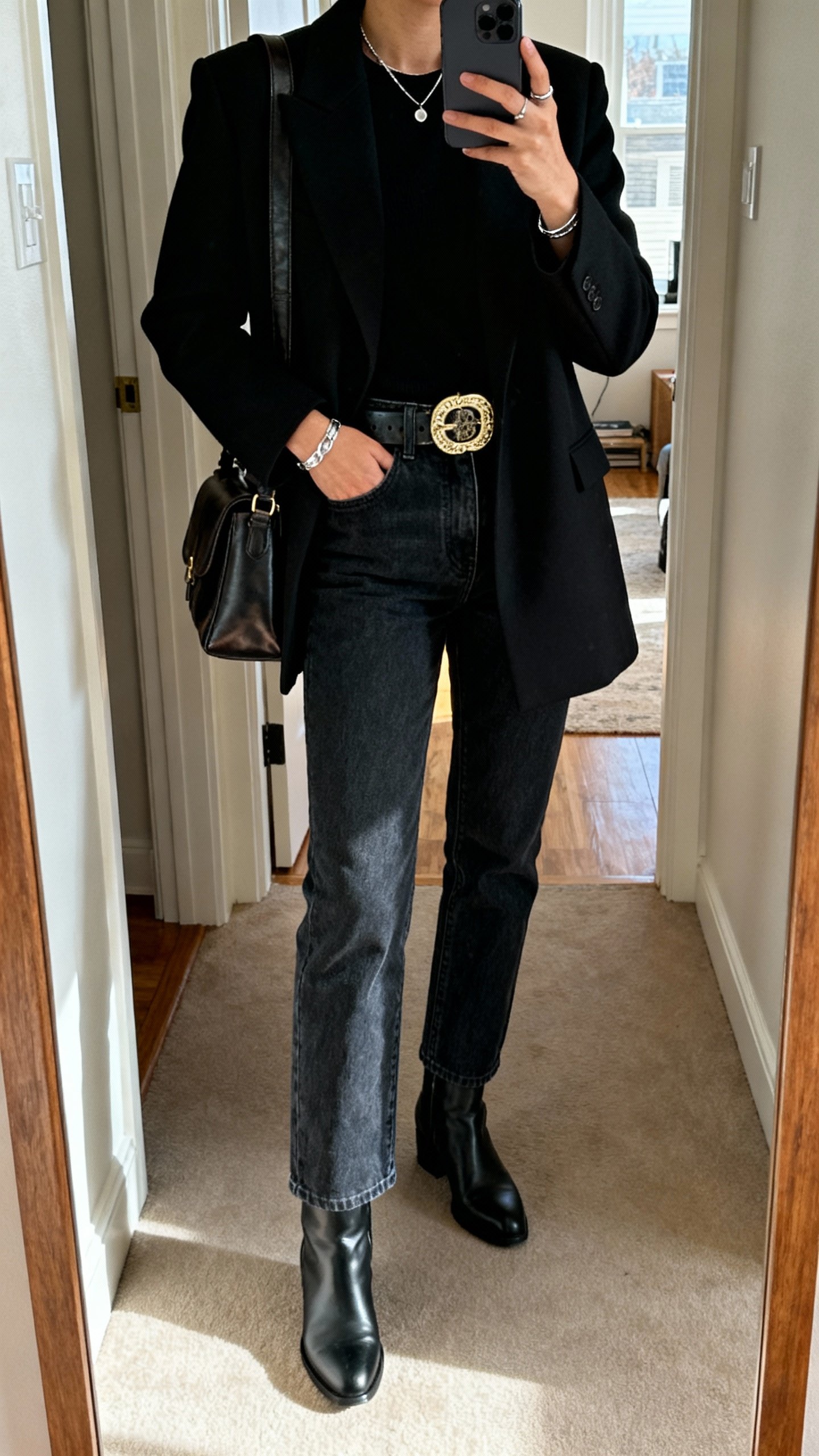 iPhone hallway mirror selfie of dark indigo straight-leg jeans with a bold statement belt, sharp black tailored coat, sleek black boots, minimal silver jewelry, leather shoulder bag, face not visible, apartment entry mirror, natural afternoon light, iPhone photo.