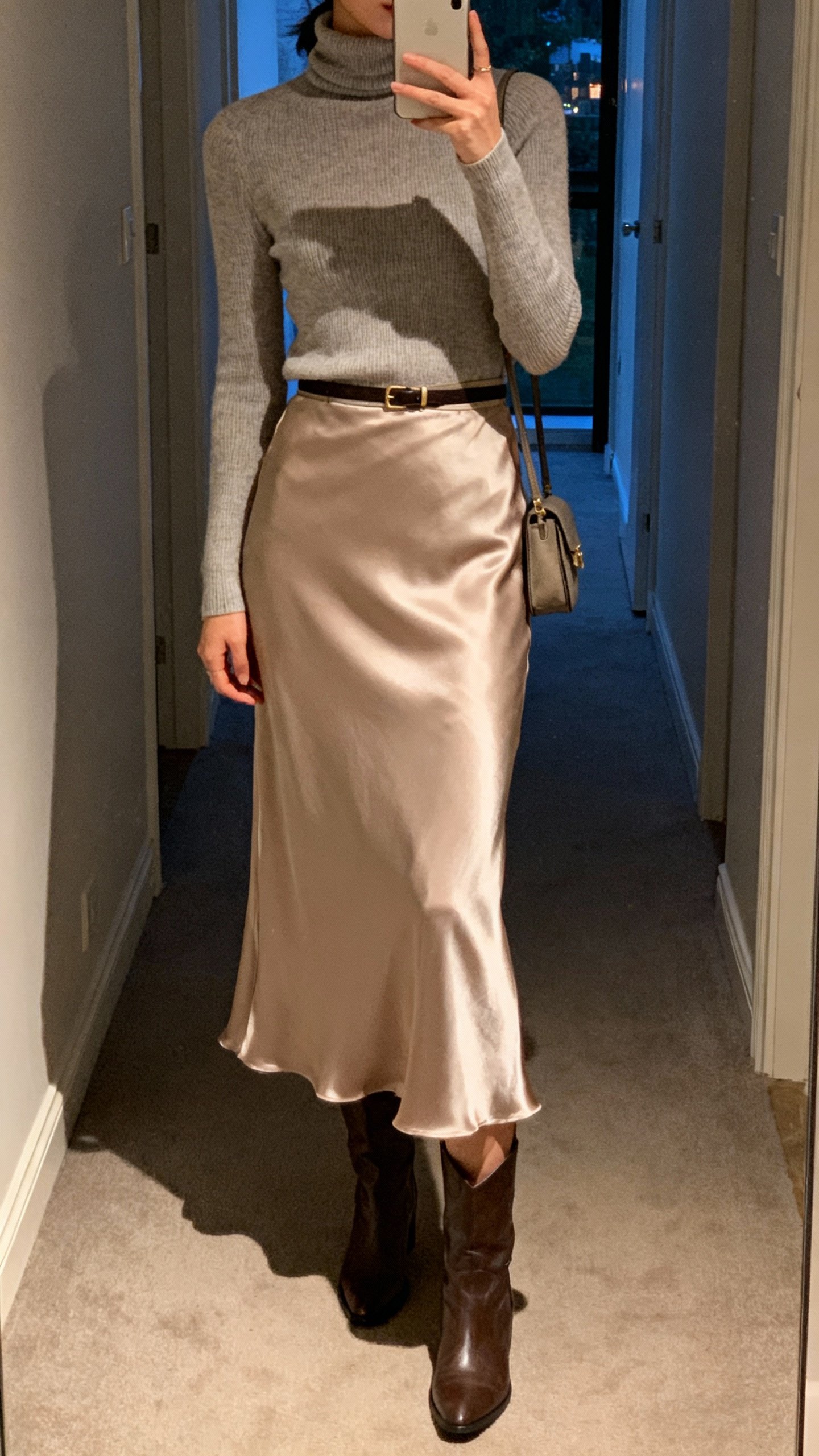 iPhone hallway selfie cropped at shoulders showing fitted turtleneck tucked into satin slip skirt with mid-calf boots, thin belt and small shoulder bag, face not visible, evening indoor light, natural iPhone photo.