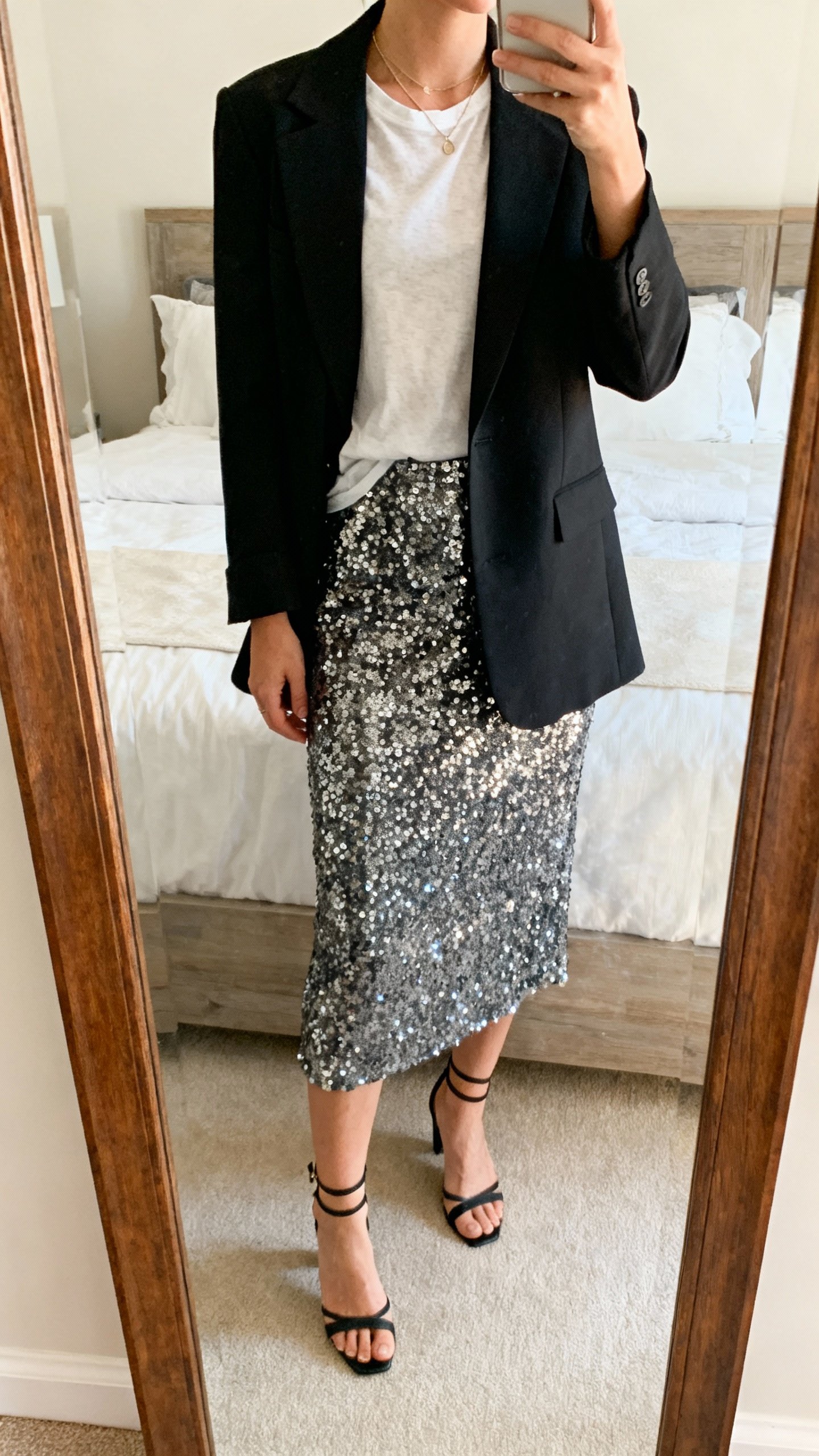 iPhone mirror selfie cropped at shoulders showing a sequin skirt with a soft tee tucked in and a structured blazer, ankle-strap heels, delicate necklace, face not visible, bedroom mirror, natural morning light, iPhone photo.