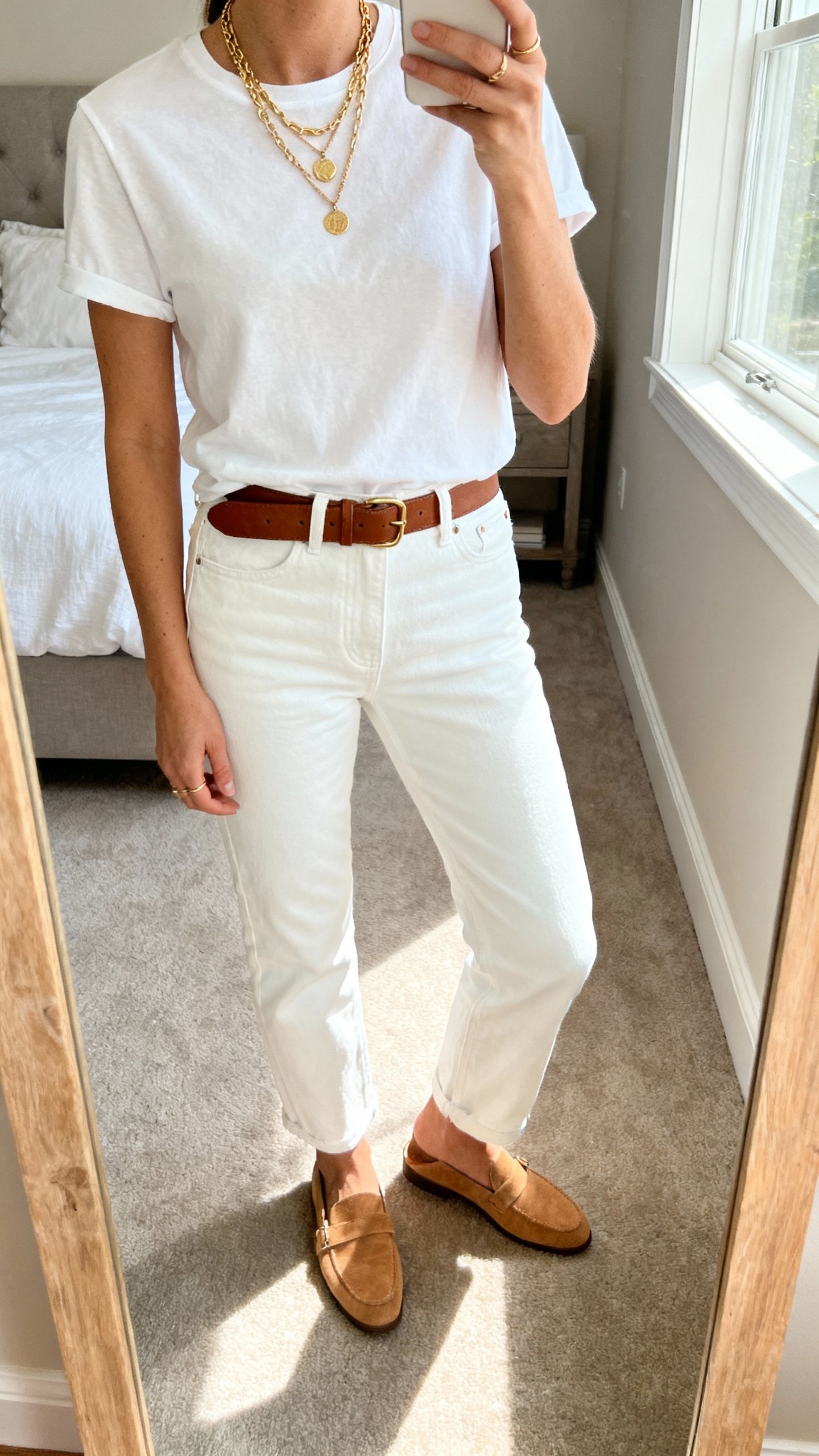 iPhone mirror selfie cropped at shoulders showing a woman in white straight-leg denim with a crisp white tee, tan loafers, layered gold necklaces, and a brown leather belt, face not visible, bedroom mirror, natural window light, iPhone photo quality.