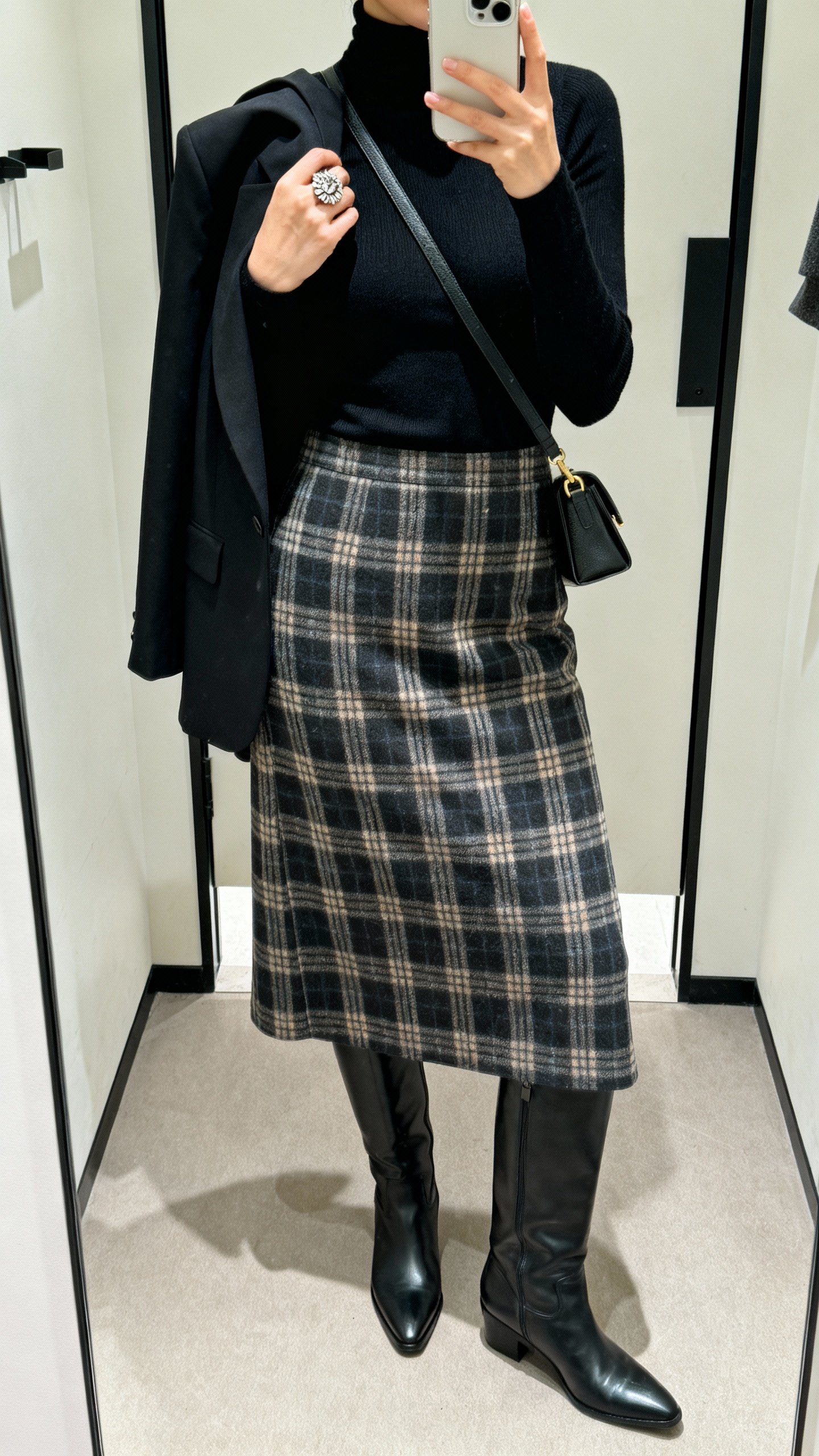 iPhone mirror selfie featuring a plaid midi skirt, fitted black turtleneck, knee-high sleek boots, structured blazer draped over shoulders, crossbody bag, statement ring, face not visible, cropped at neck, boutique fitting-room lighting, natural iPhone photo.