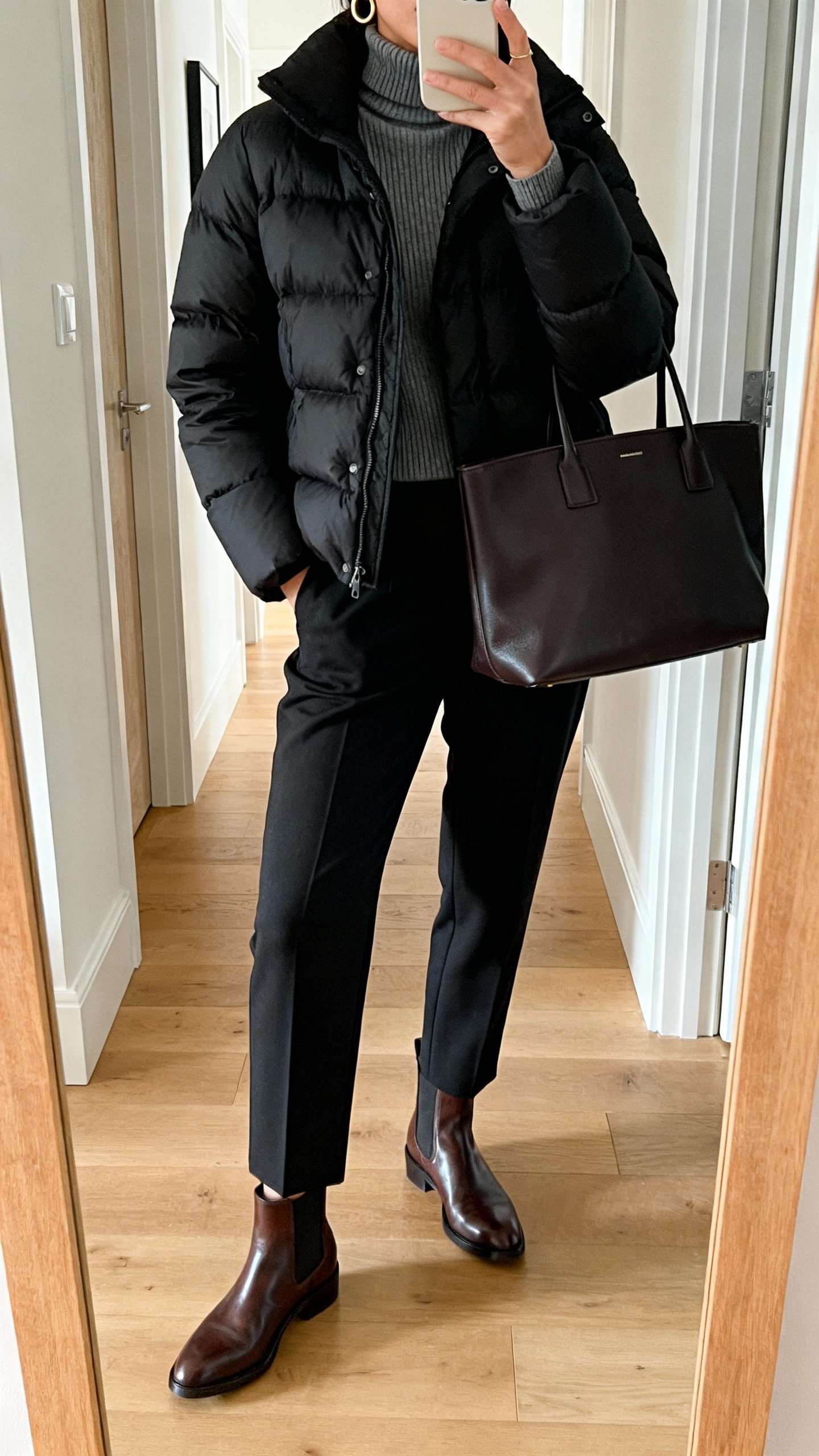 iPhone mirror selfie in entryway: sleek black puffer jacket styled with tapered trousers, fine-gauge turtleneck, leather Chelsea boots, structured tote, subtle gold hoops, face not visible, cropped at neck, natural morning light, casual iPhone photo.