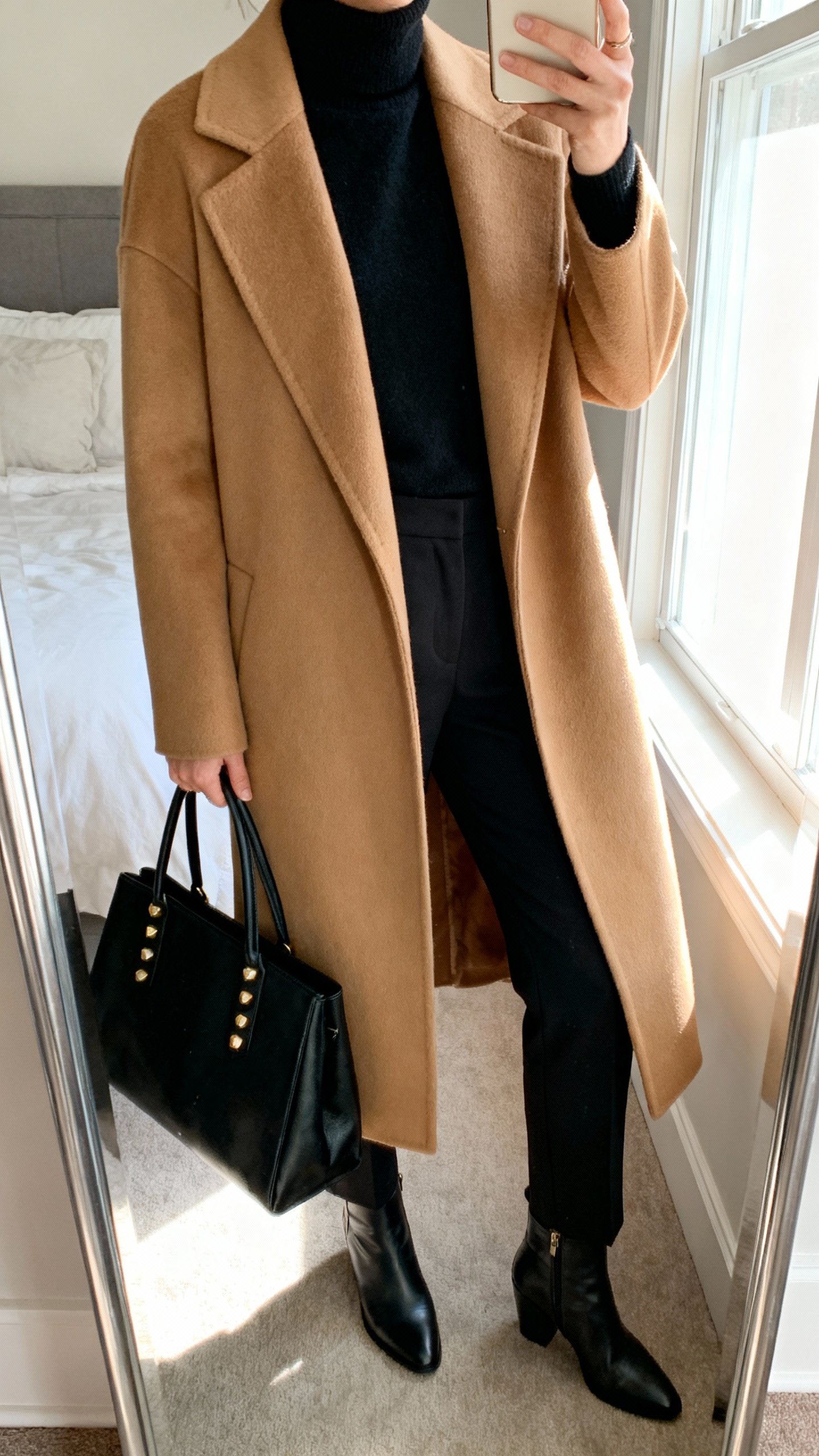 iPhone mirror selfie of a luxe camel coat over a monochrome black base (black turtleneck, black tailored trousers), sleek ankle boots, simple gold studs, structured black tote, face not visible, cropped at neck, bedroom mirror with window light, natural iPhone photo quality.