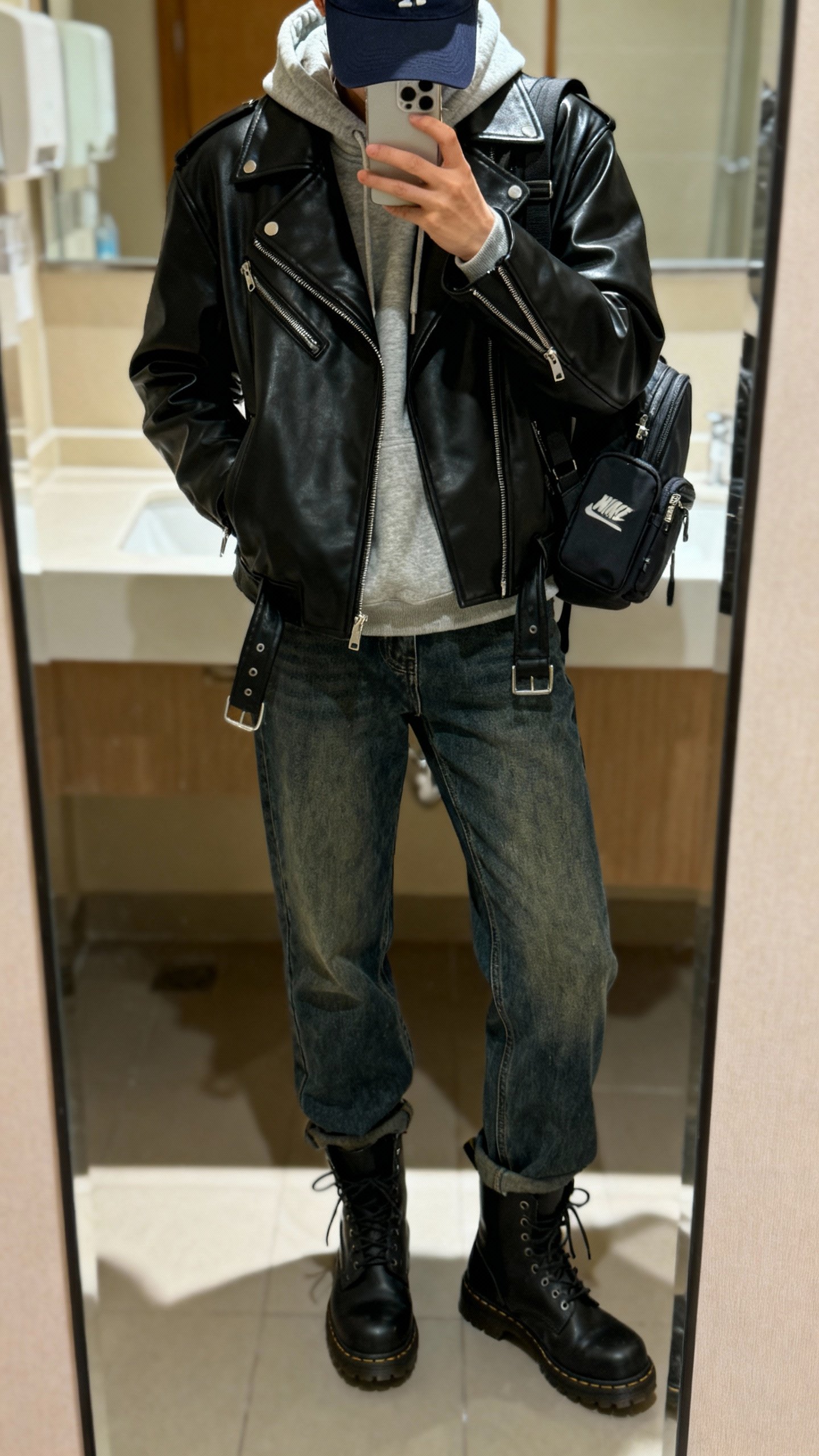iPhone mirror selfie of black leather moto jacket layered over cozy hoodie with straight jeans and combat boots, baseball cap and mini backpack, face not visible, bathroom mirror, indoor lighting, casual iPhone quality.