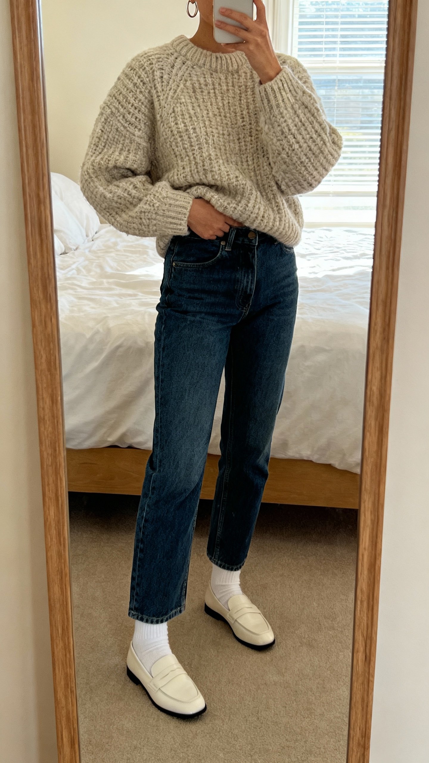 iPhone mirror selfie of chunky knit sweater half-tucked into straight-leg jeans with white socks and loafers, simple hoop earrings, face not visible, cropped at neck, bedroom full-length mirror, soft morning window light, natural iPhone photo quality.