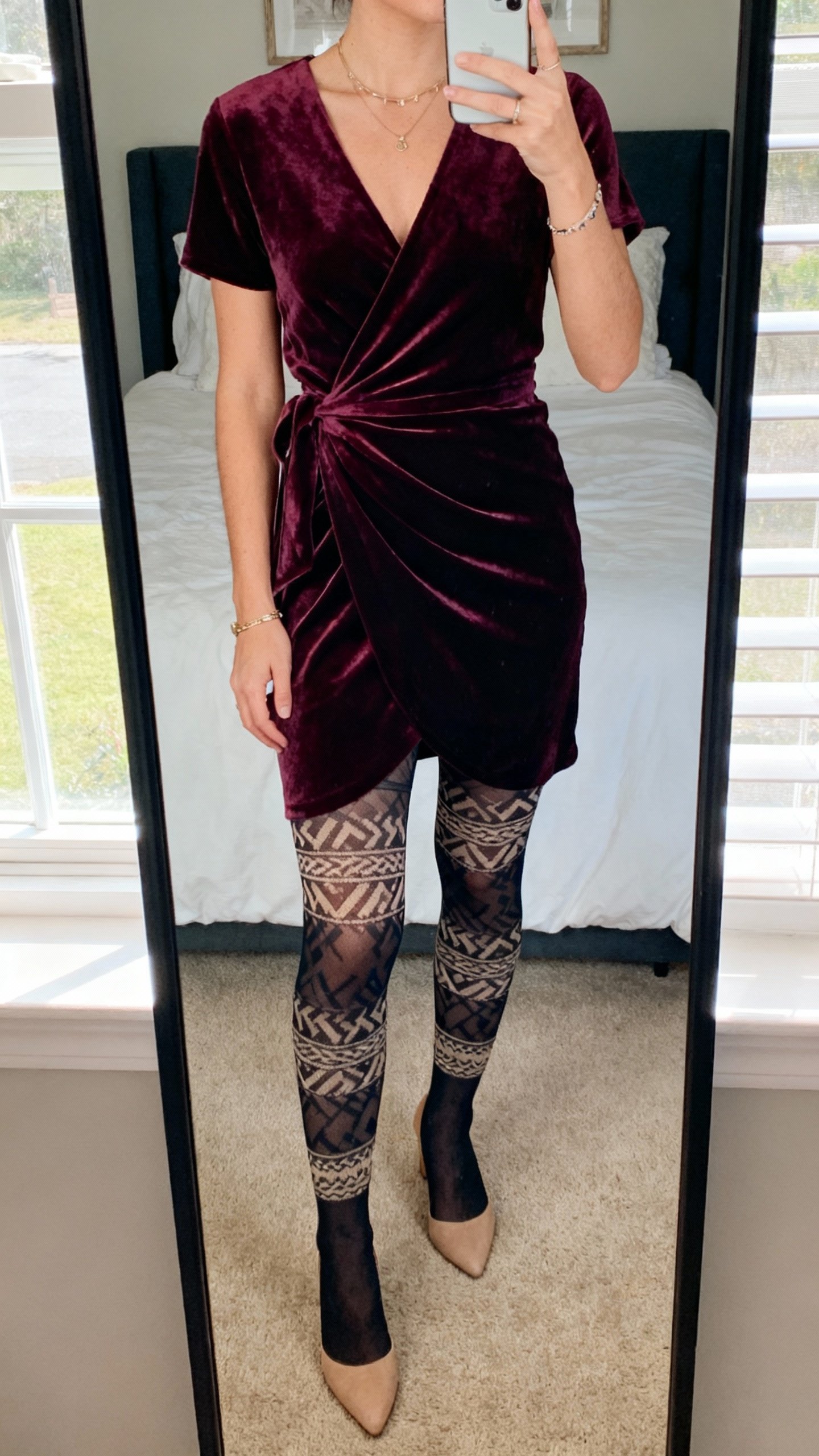 iPhone mirror selfie showing a velvet wrap dress with statement patterned tights and simple heels, minimal jewelry, face not visible, cropped at neck, bedroom mirror with soft window light, natural iPhone photo quality.