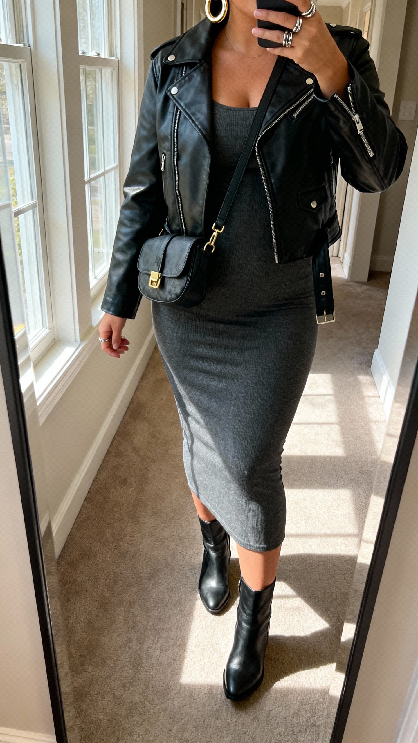 iPhone mirror selfie showing curve-hugging midi dress, black leather moto jacket, ankle boots, crossbody bag, chunky hoops and stacked rings, face not visible, cropped at neck, hallway mirror with morning window light, natural iPhone photo quality