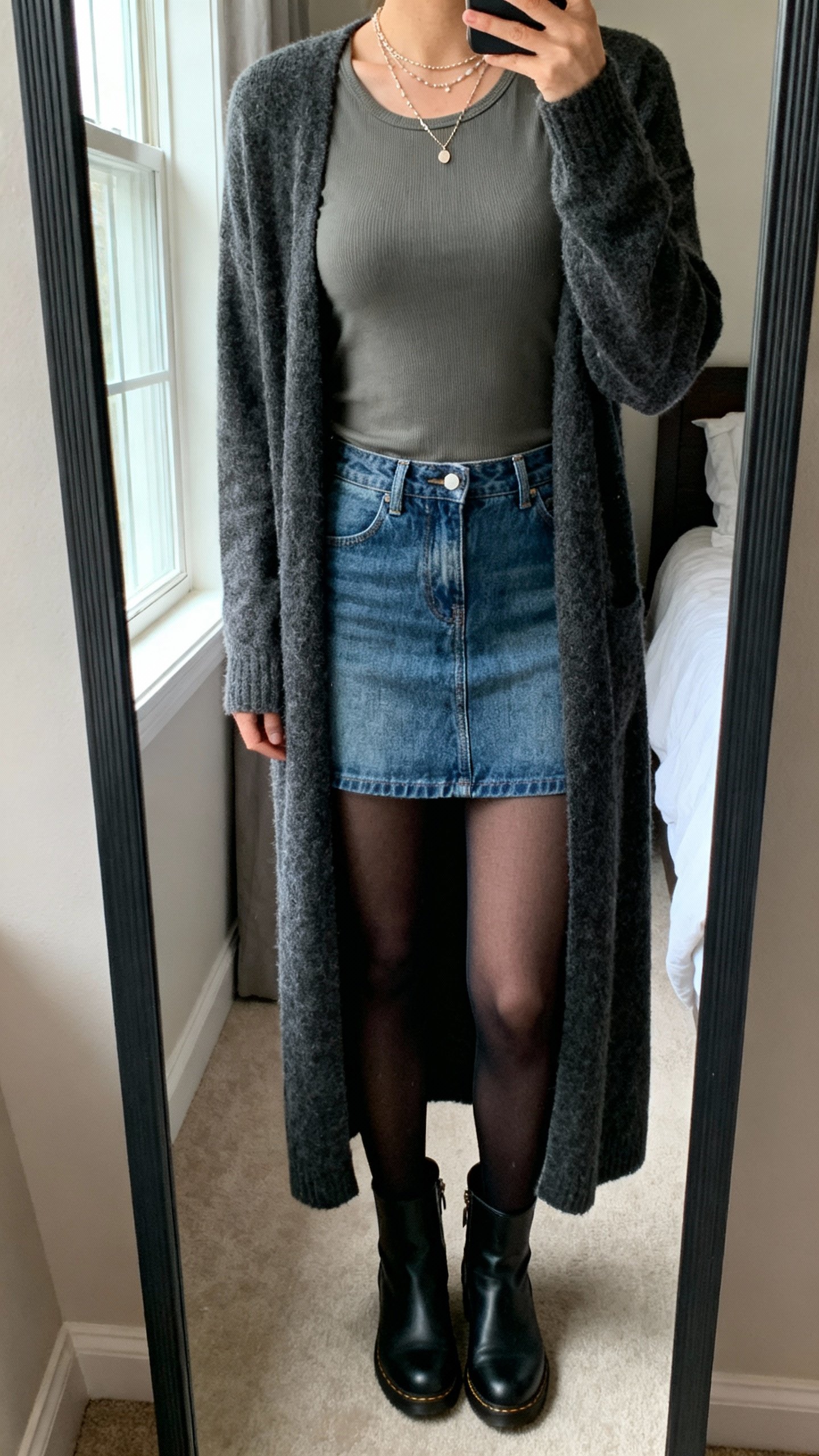 iPhone mirror selfie showing longline cardigan over fitted tee with denim skirt, opaque tights, ankle boots, layered delicate necklaces, face not visible, cropped at neck, bedroom mirror, soft window light, natural iPhone photo quality.