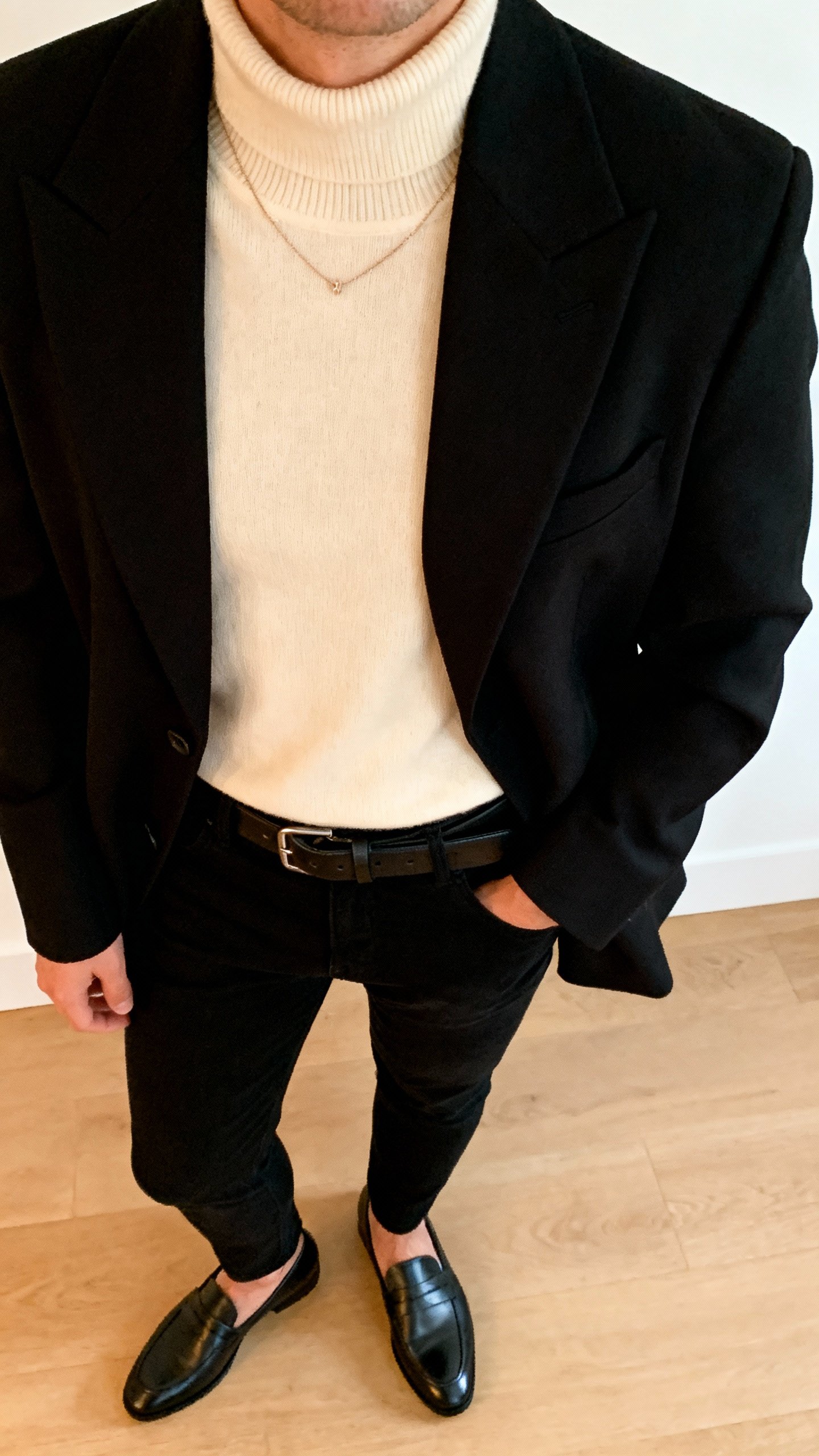 iPhone office-bathroom mirror selfie of a structured black blazer layered over a cream turtleneck, slim black pants, sleek loafers, leather belt, dainty necklace, face not visible, cropped at neck, warm indoor lighting, casual iPhone quality.
