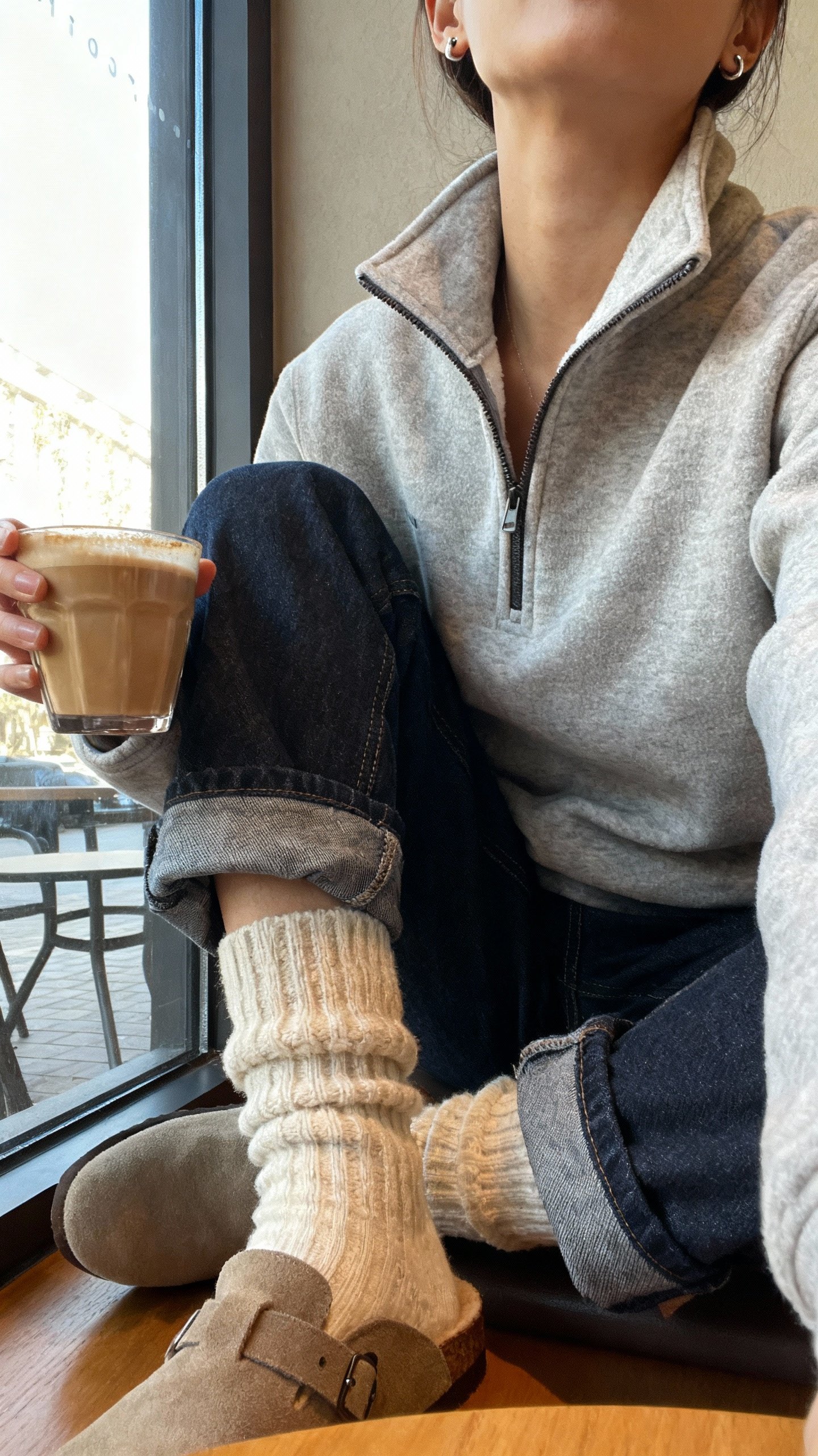 iPhone seated cafe selfie cropped at neck: half-zip fleece tucked slightly into barrel jeans with chunky socks and mules, simple studs and latte in hand, face not visible, window light, natural iPhone aesthetic.